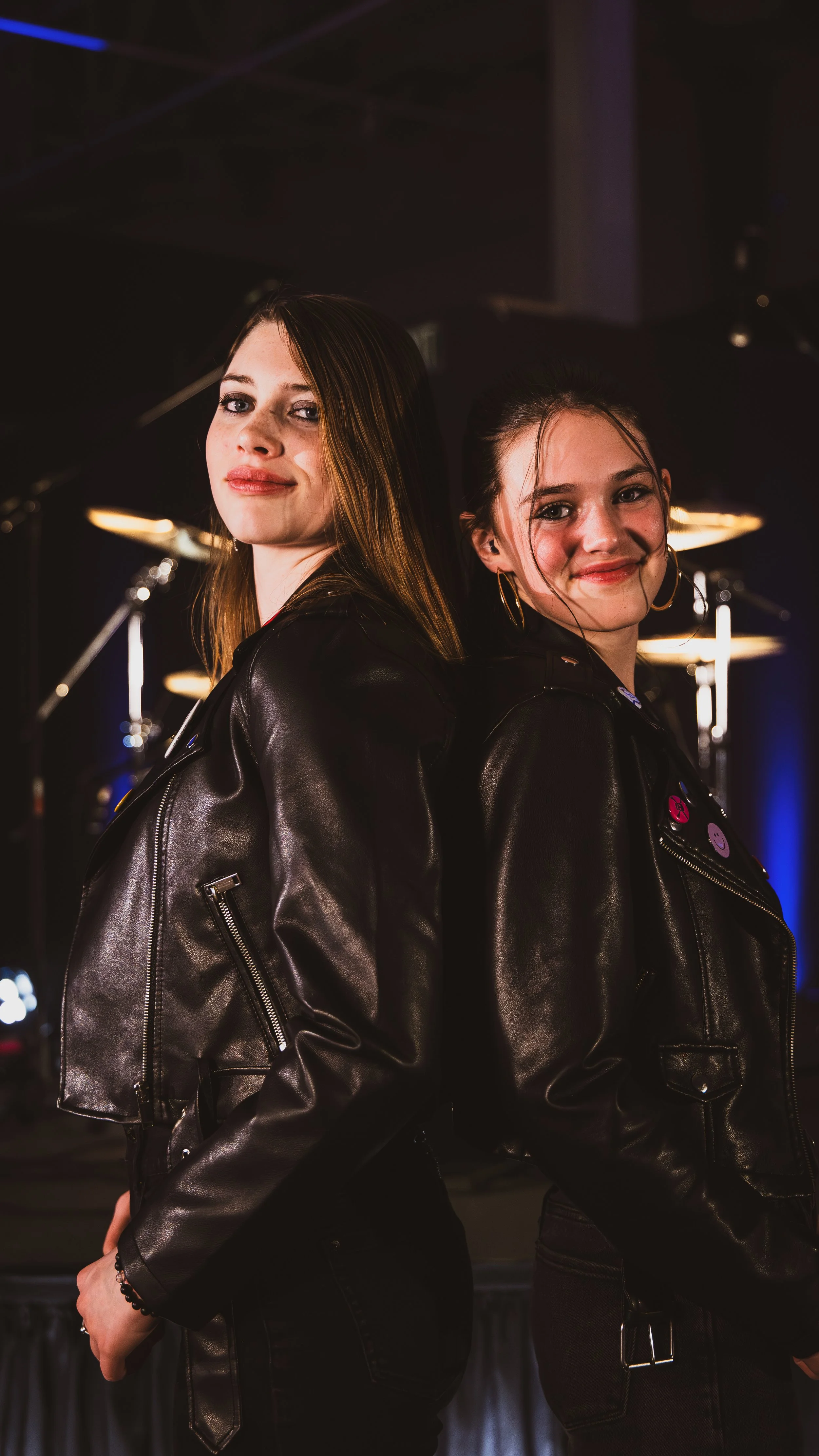 Two young women standing back to back in a dark room with stage lighting, wearing black leather jackets and black pants, smiling at the camera.