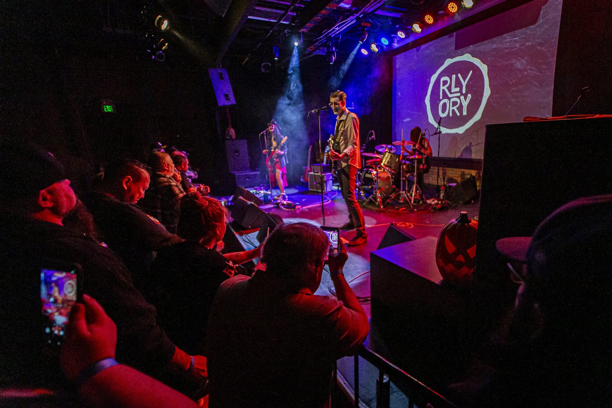 A band performing on stage at an event called RLY RY. The stage has purple and red lighting, with a singer and at least two other musicians. Audience members are seated and watching the performance, some capturing photos with their phones. The backgr