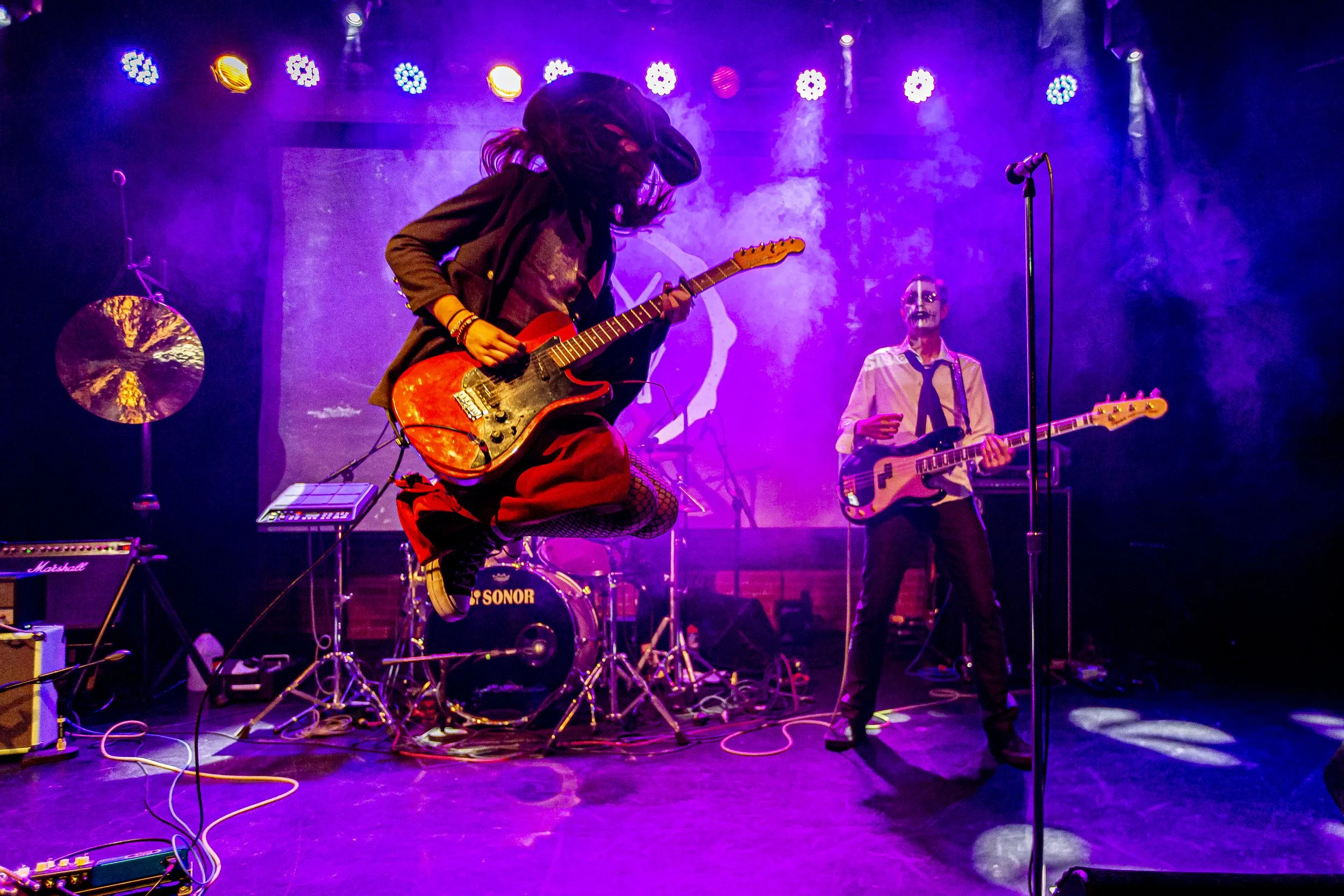 A band performing on stage with a guitarist jumping in the air. The stage is lit with colorful lights, and two other band members are playing guitars. There are various musical equipment and cables on the stage.