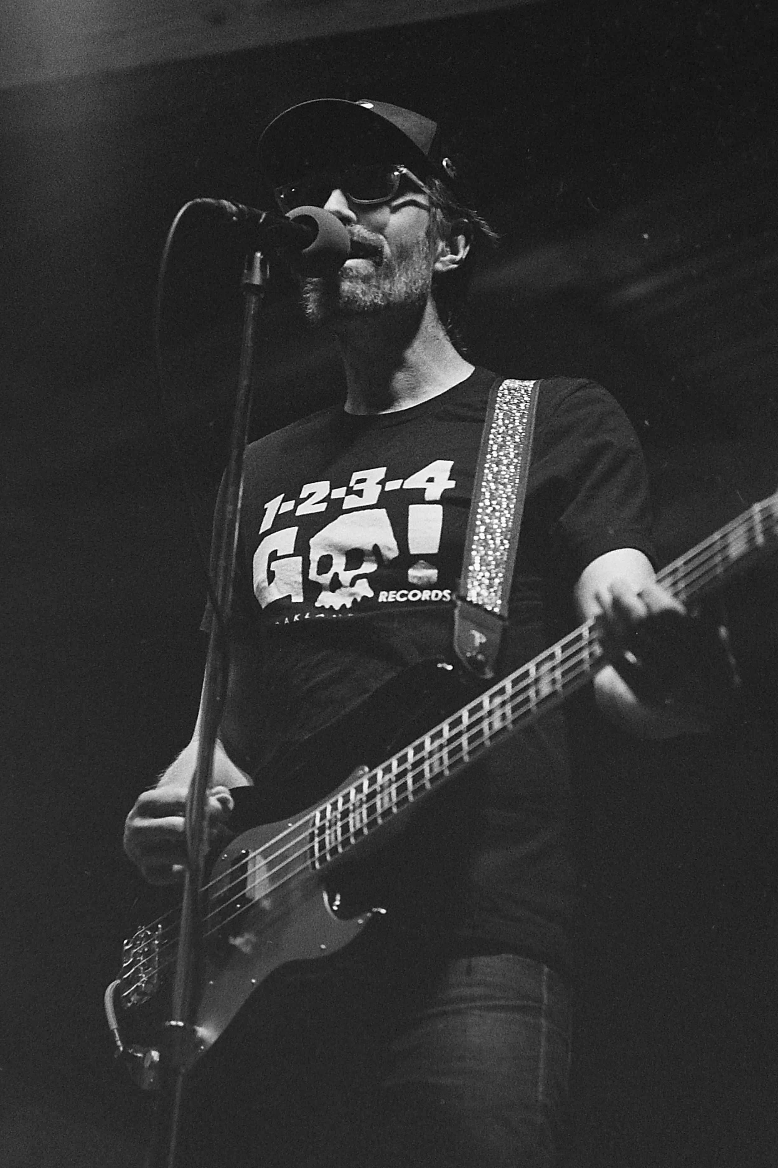 A man with glasses and a hat singing into a microphone while playing an electric bass.
