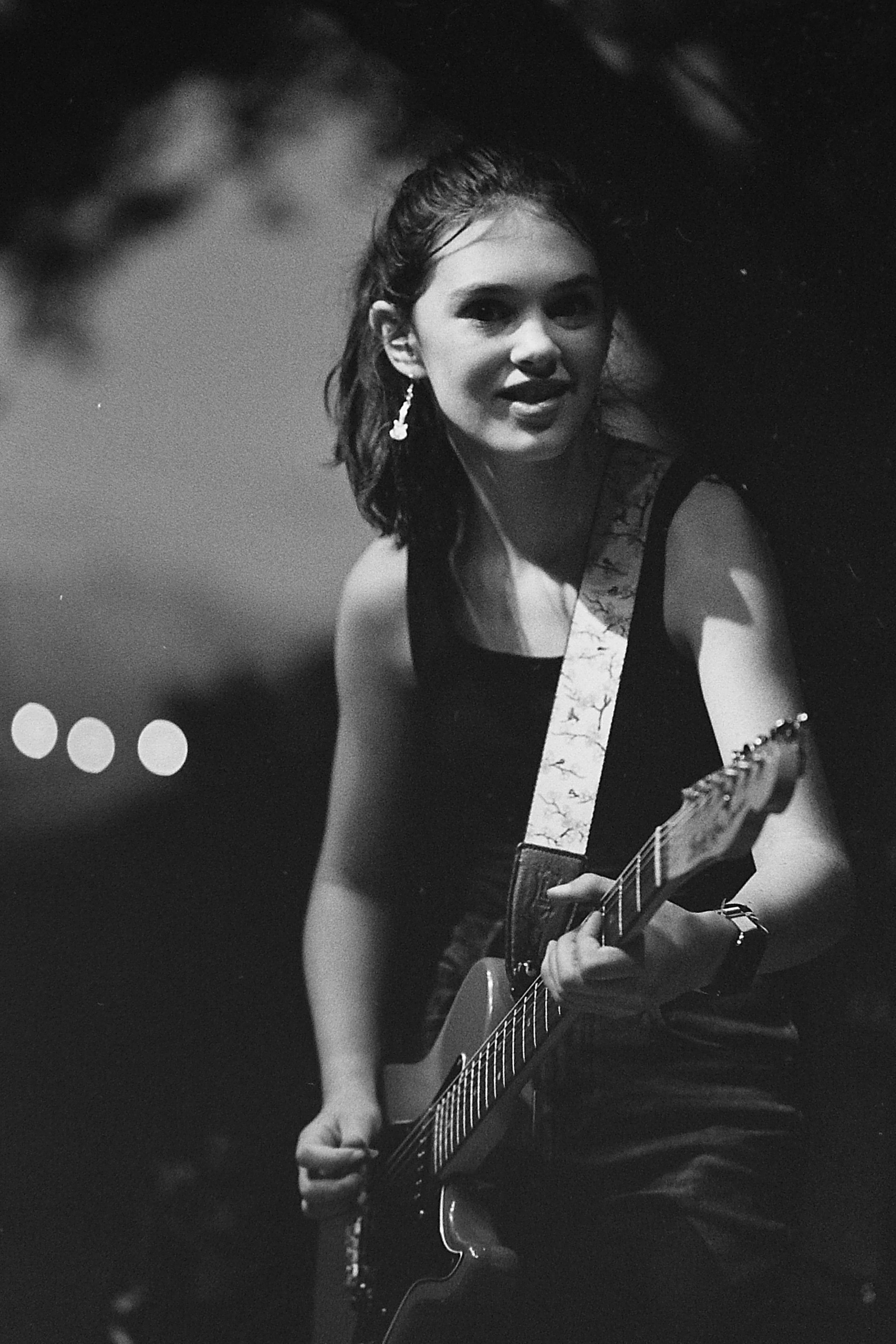 Black and white photo of a woman playing an electric guitar. She has short, wavy hair and is wearing earrings and a sleeveless top. She appears to be performing or practicing, with a focused expression.