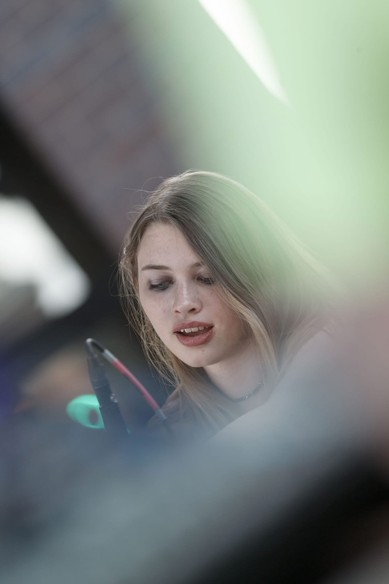 A young woman with light brown hair looking at something in front of her, possibly a phone, with a blurred background and a cloudy, outdoors setting.