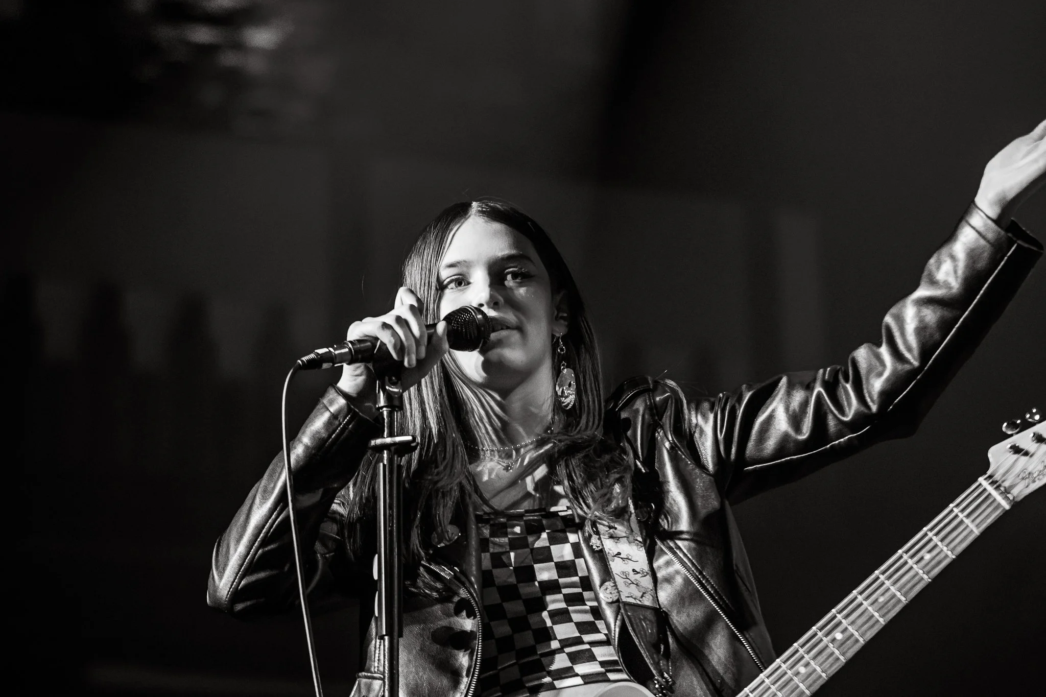 A woman with long hair singing into a microphone on stage, wearing a leather jacket and holding a guitar.