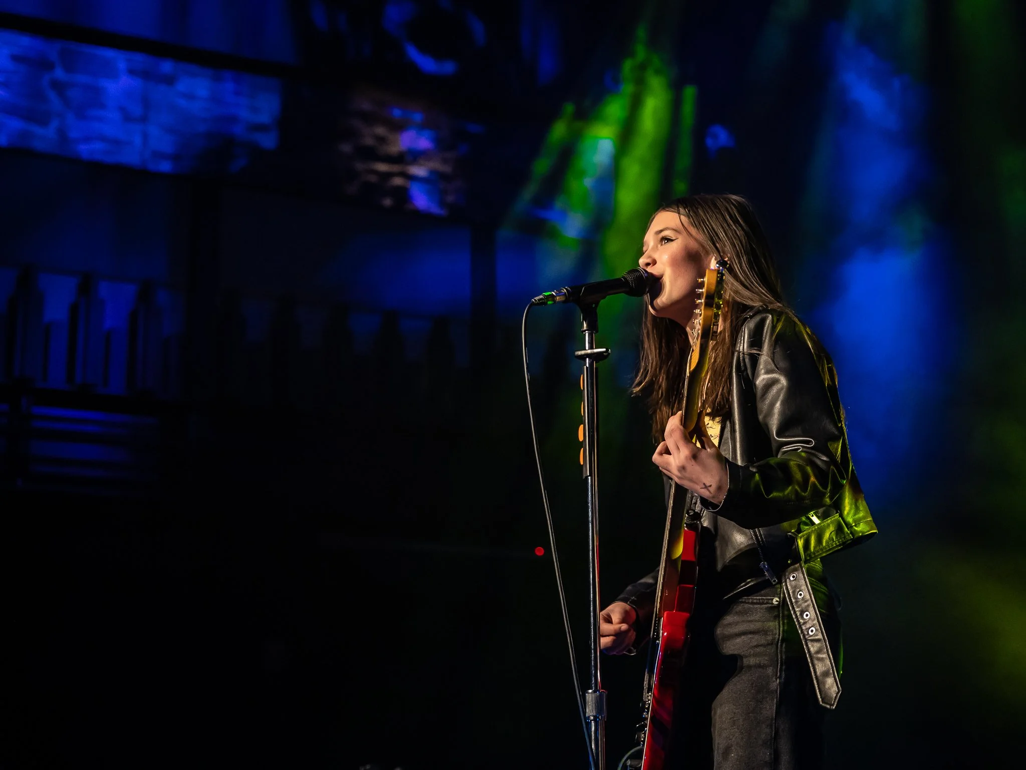 A woman with long brown hair singing into a microphone on stage, holding a guitar, wearing a leather jacket, with colorful stage lighting and a dark background.