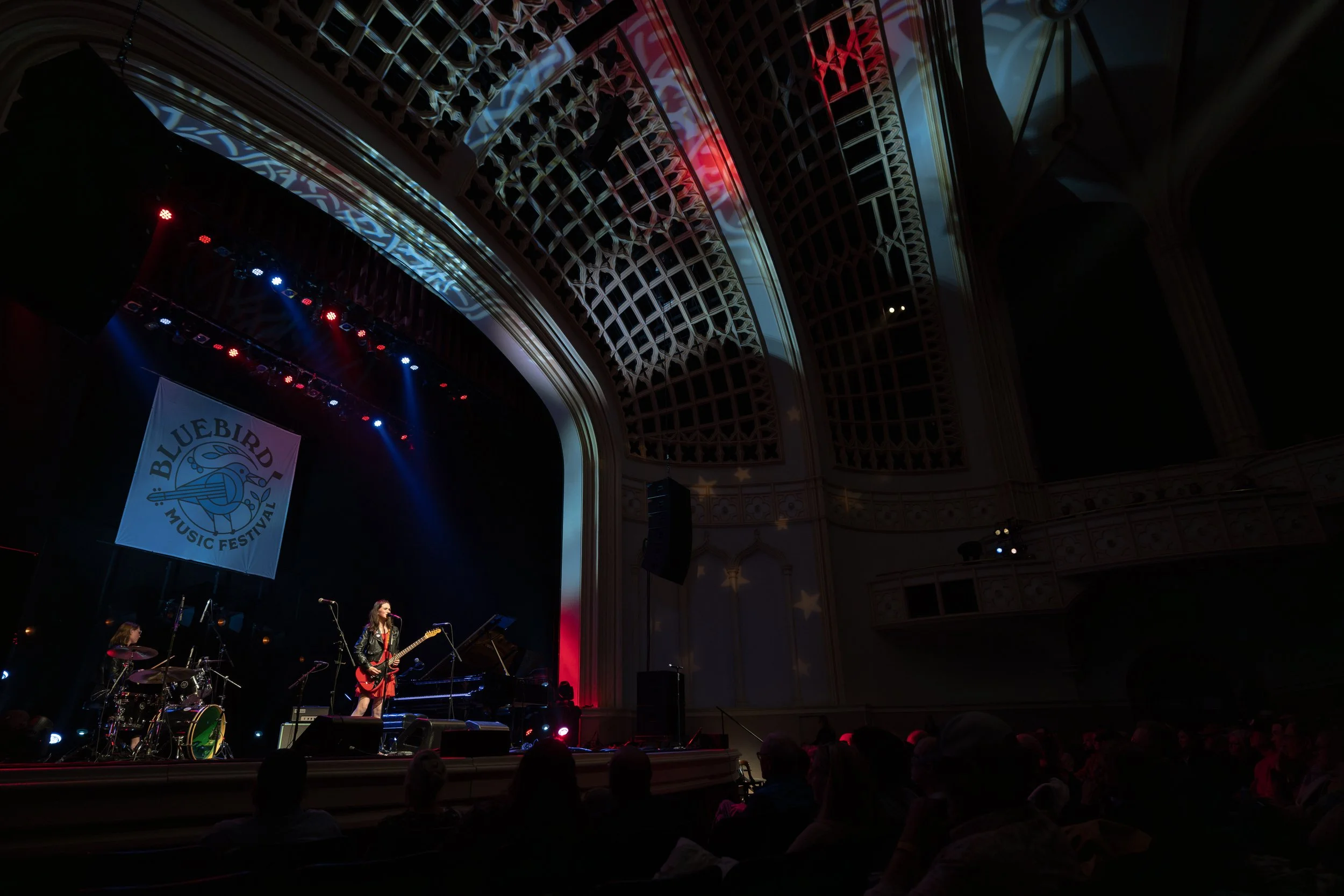 A live music performance on stage at the Bluebird Music Festival, with band members playing instruments and a backdrop featuring the festival's logo, inside a grand hall with ornate architectural details and colored lighting.