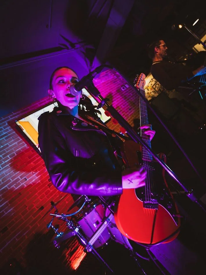 A female musician singing into a microphone and playing an acoustic guitar during a live performance, with a male musician playing keyboard in the background, in a dimly lit venue with purple and red lighting.