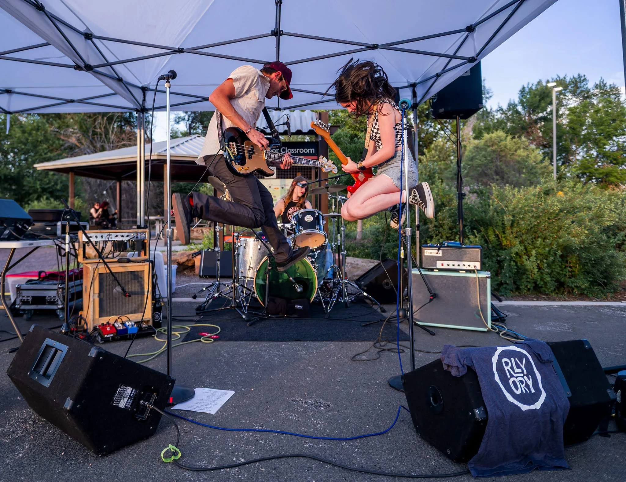 Two musicians performing on an outdoor stage under a canopy, one playing an electric bass guitar and the other playing a guitar, with a drummer in the background. Equipment and instruments are scattered on stage, and there is a sign that reads 'for c