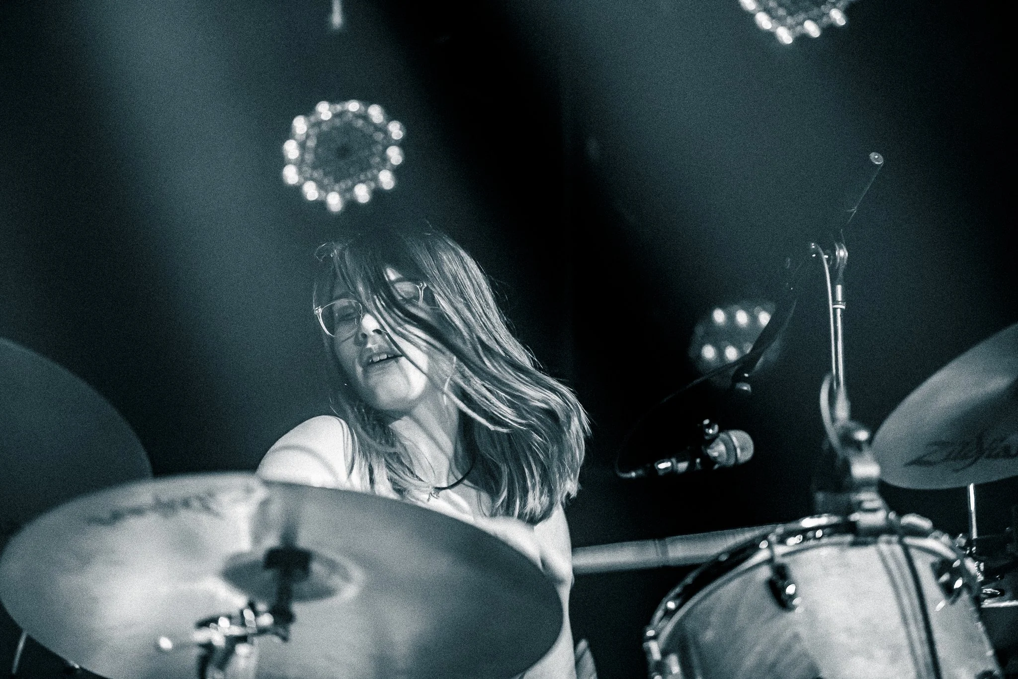 A woman with long hair and glasses playing drums in a dark setting with stage lights.