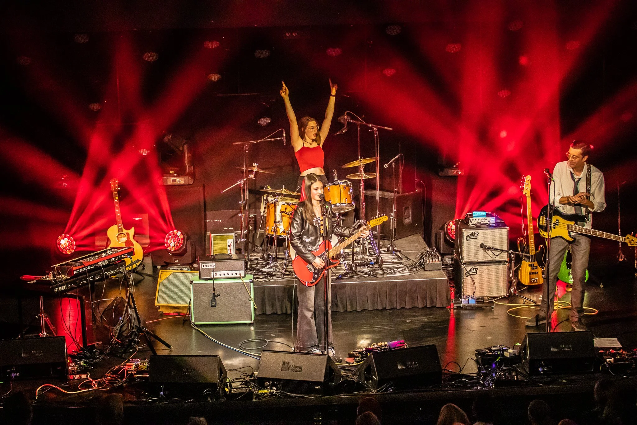 Band performing on stage with guitars, drums, and microphones, red stage lighting, and audience in the foreground.