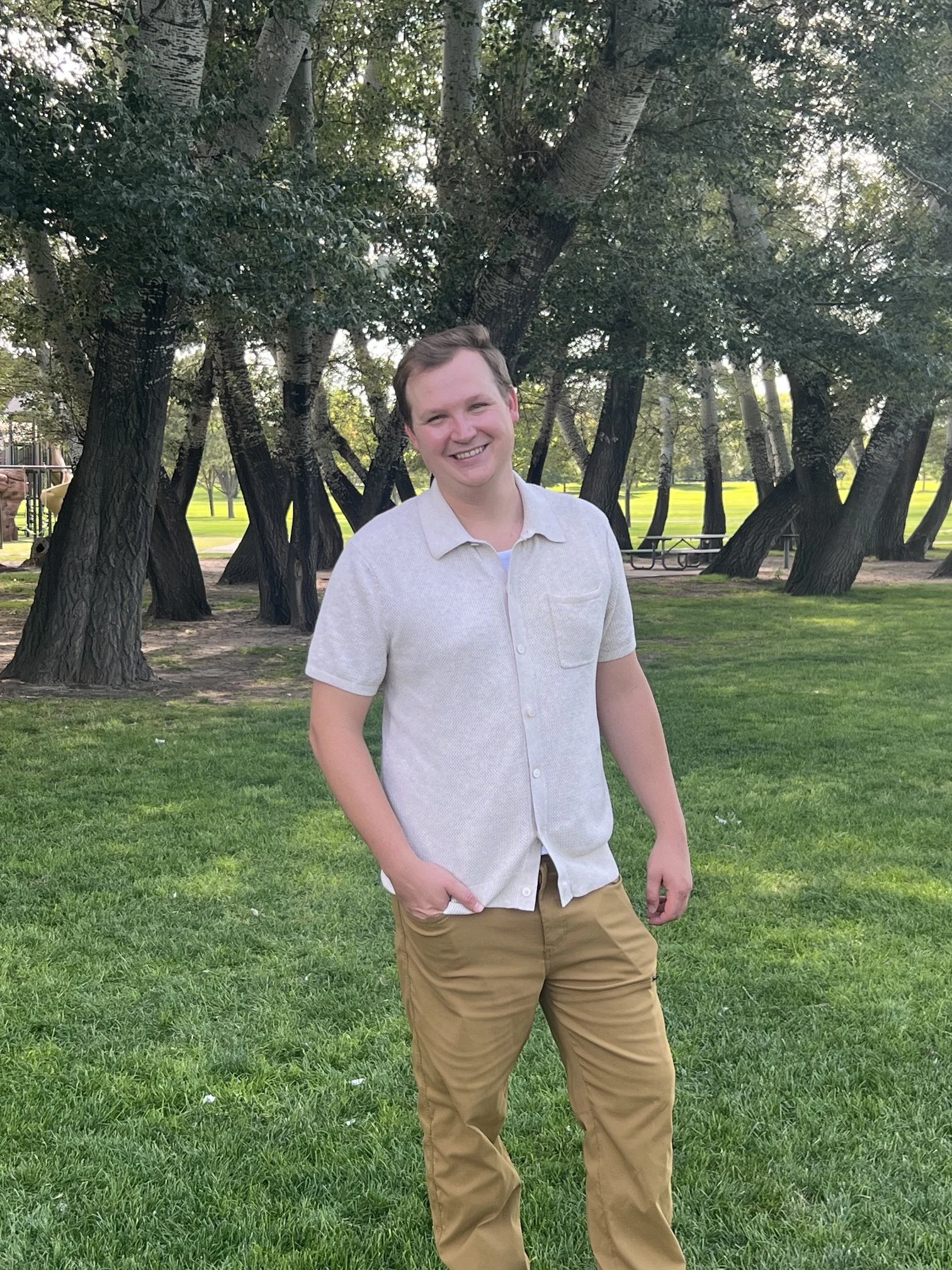 A young man with short hair smiling, standing outdoors in a park with green grass and trees in the background.