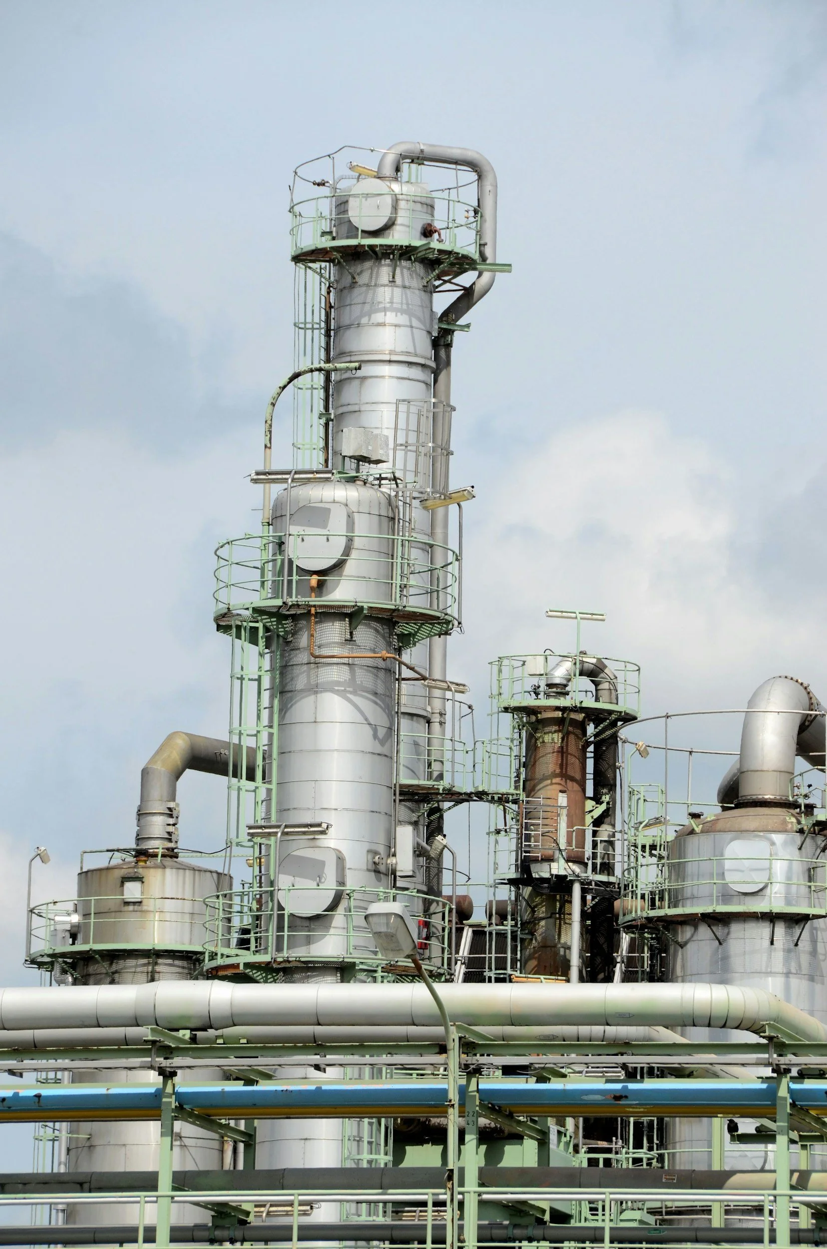 An industrial factory with multiple metal pipes, tanks, and walkways against a cloudy sky.