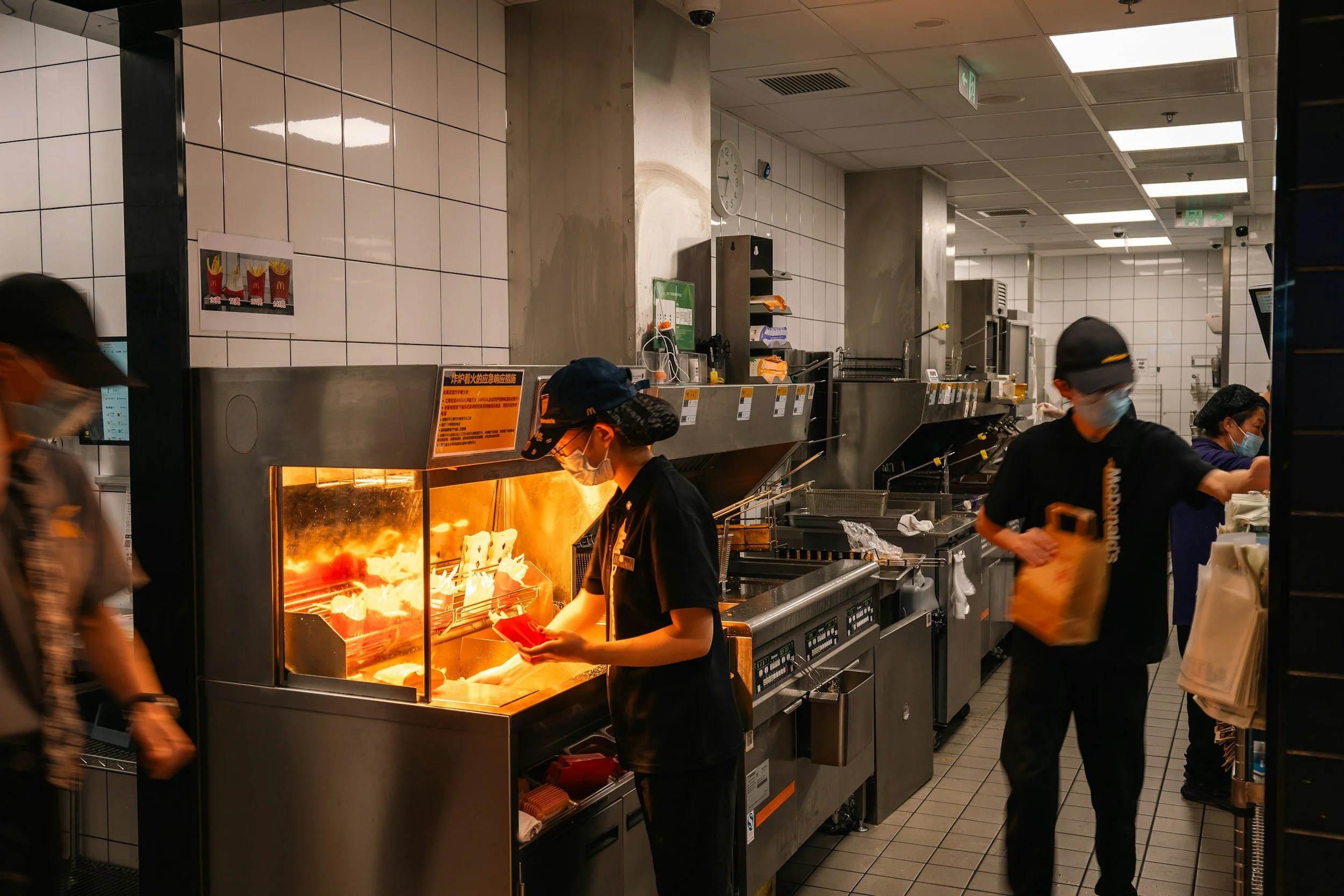 Fast food restaurant kitchen with employees preparing food, including a worker near a hot grill and others working at counters, with tiled walls and overhead lighting.