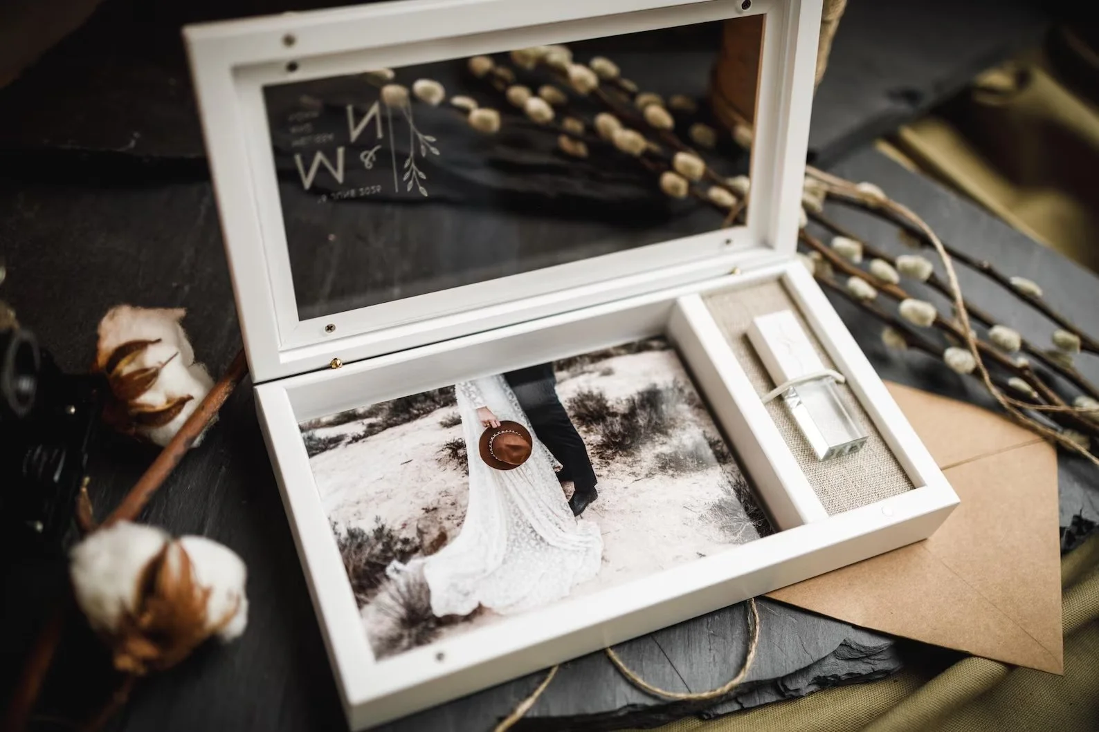 Una scatola bianca con uno specchio aperta contiene una foto di una coppia in abiti da sposa e da sposo in un paesaggio naturale, vicino a rami di bacche e oggetti decorativi rustic.