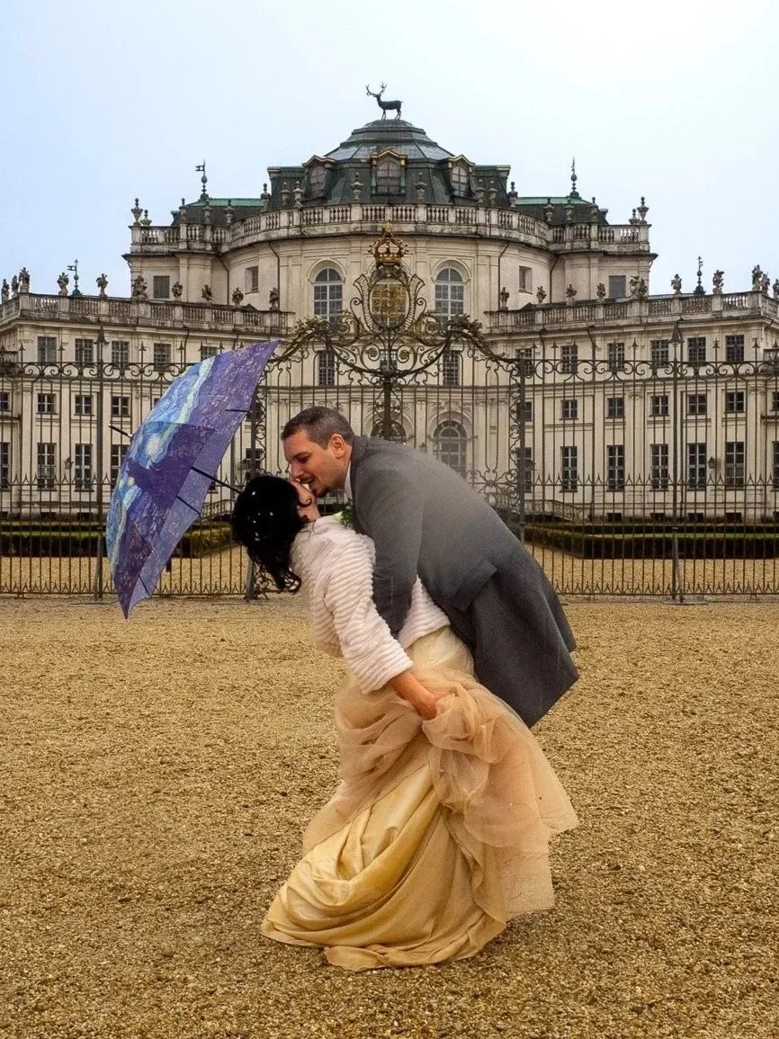 Una coppia felice, un uomo e una donna, sotto un ombrello colorato, si abbracciano e sorridono di fronte a un grande edificio antico con un'escrescenza di cervo sulla cupola, in una giornata di pioggia.