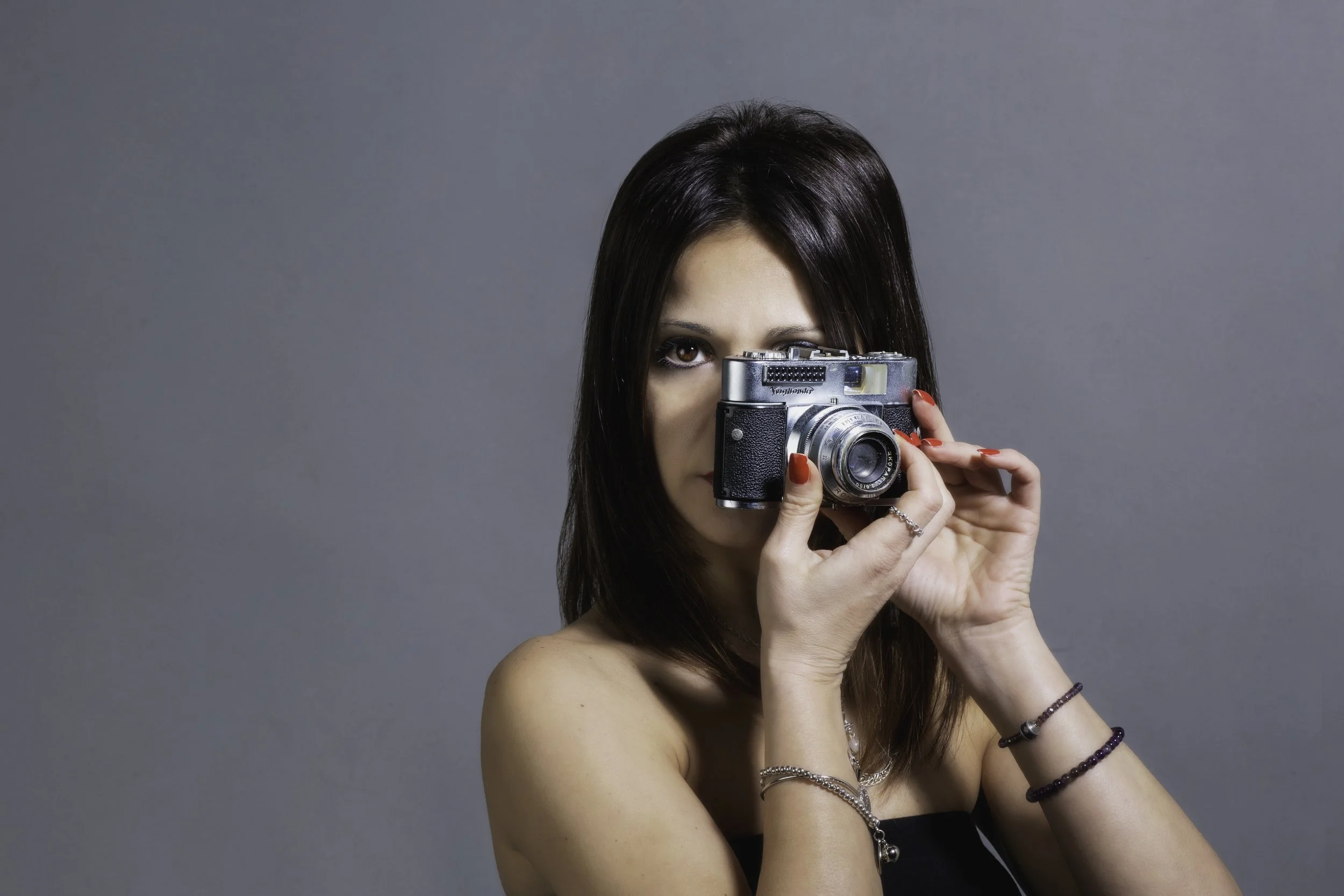 Una donna con i capelli castani scuri si sta pelando con una macchina fotografica vintage. Indossa numerosi braccialetti e mostra unghie rosse.