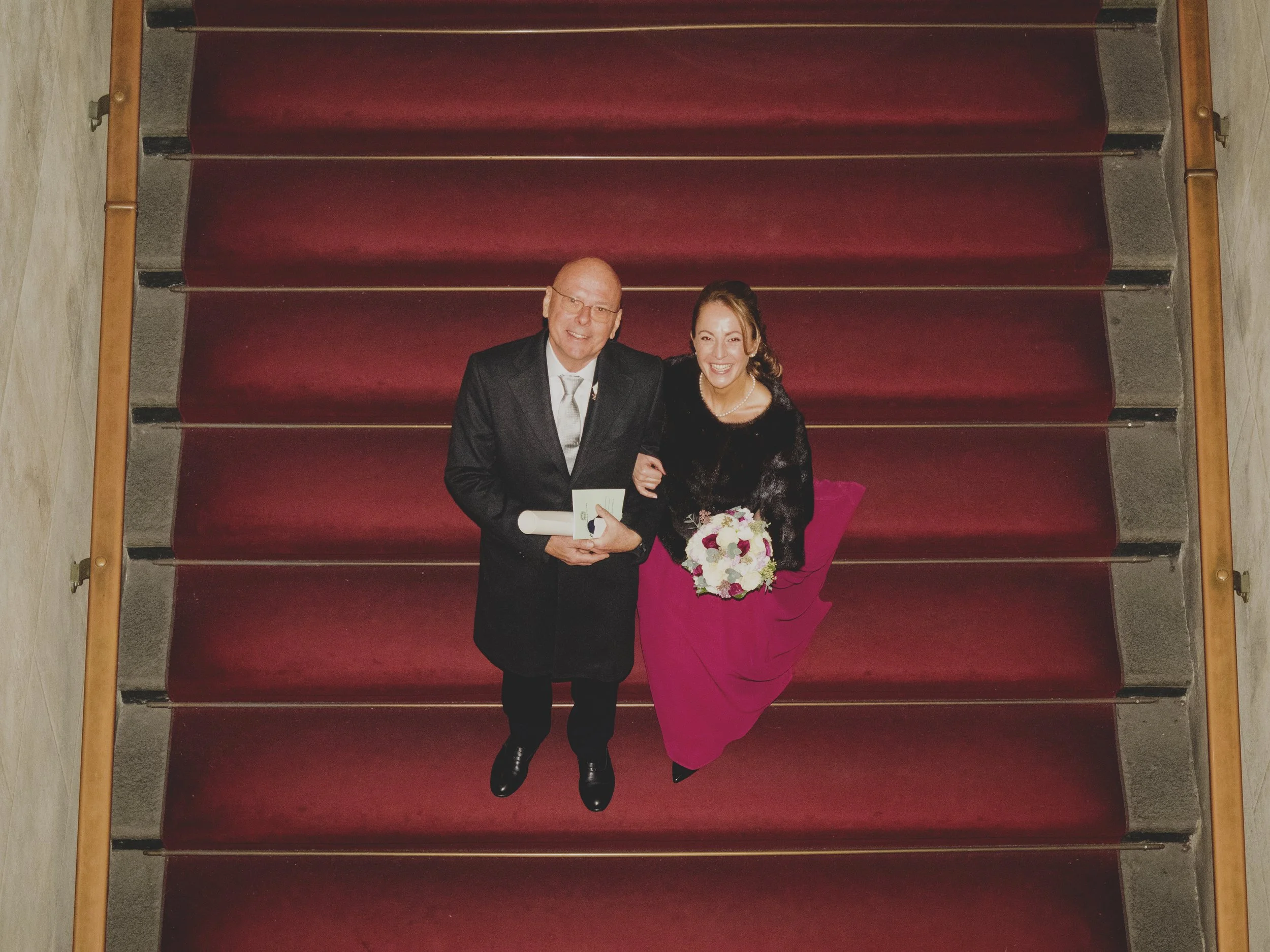 Uomo elegante con giacca e cravatta e donna in abito rosa con bouquet di fiori, entrambi sorridenti, in piedi su una scaletta ricoperta di tappeto rosso.
