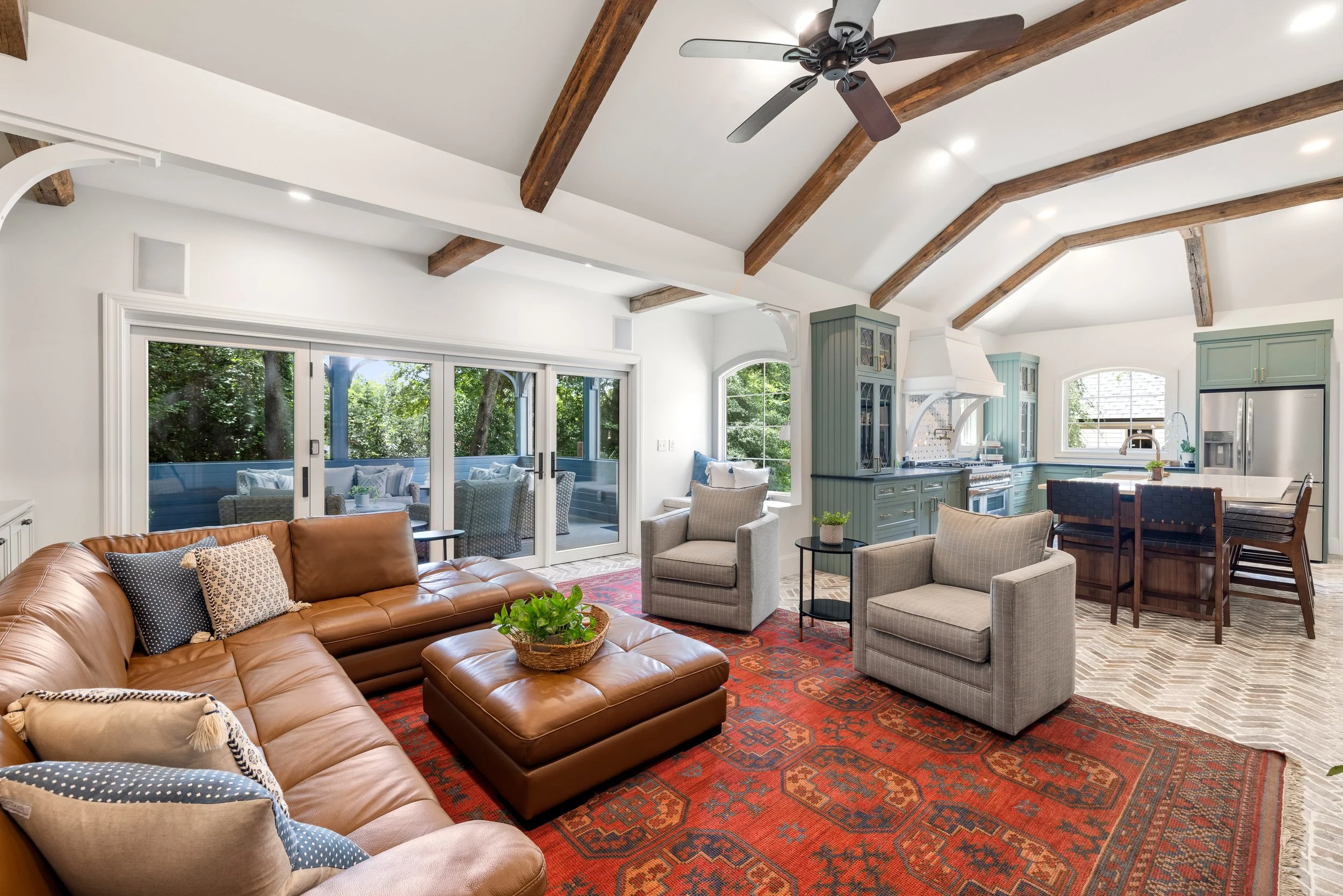 Living room with vaulted ceiling, wooden beams, ceiling fan, large windows, sliding glass doors, beige and gray seating, leather couch, patterned rugs, and open kitchen with mint green cabinets.