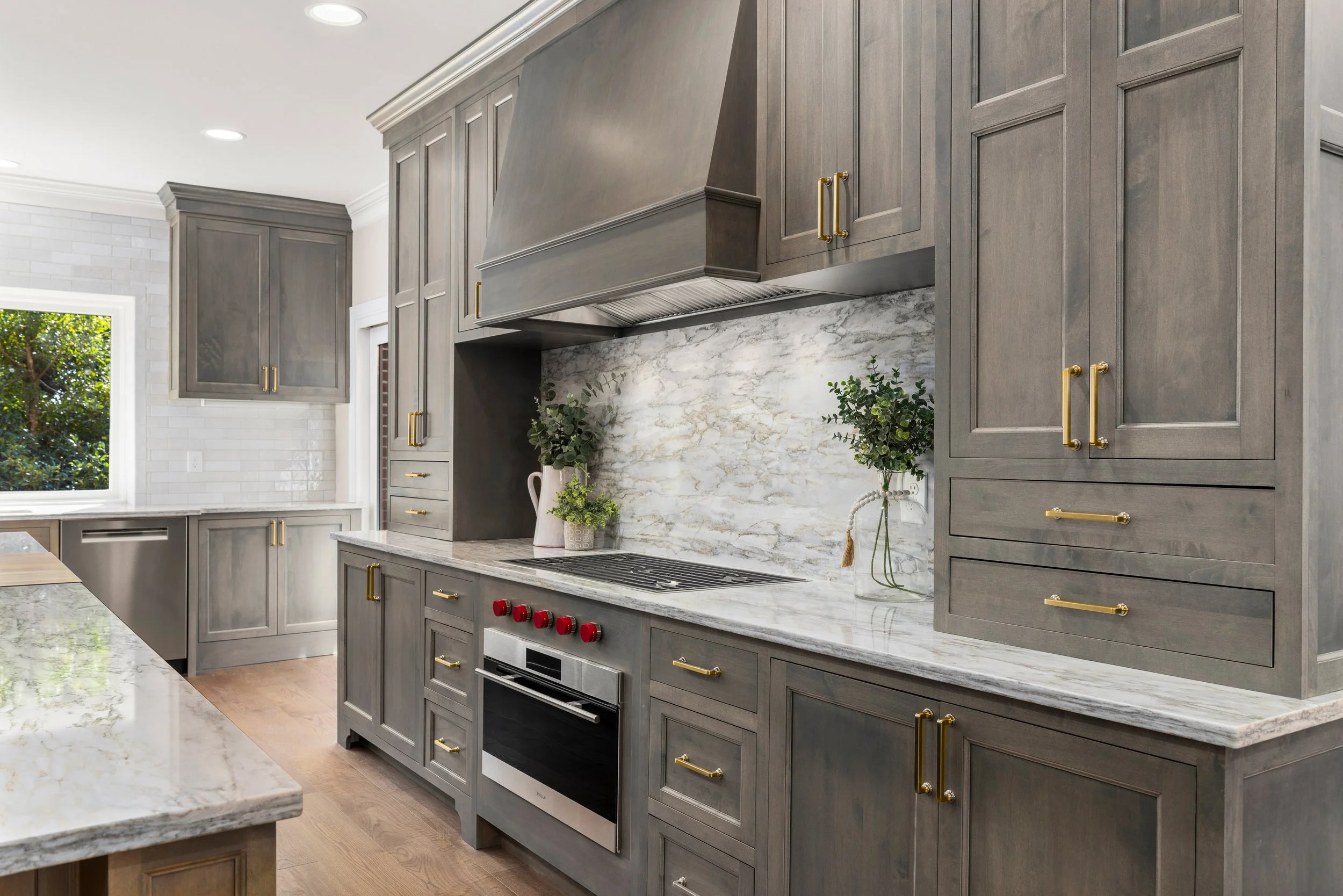 Modern kitchen with gray cabinetry, marble countertops, and a marble backsplash. Includes a stove with red knobs, decorative plants, and a window showing greenery outside.