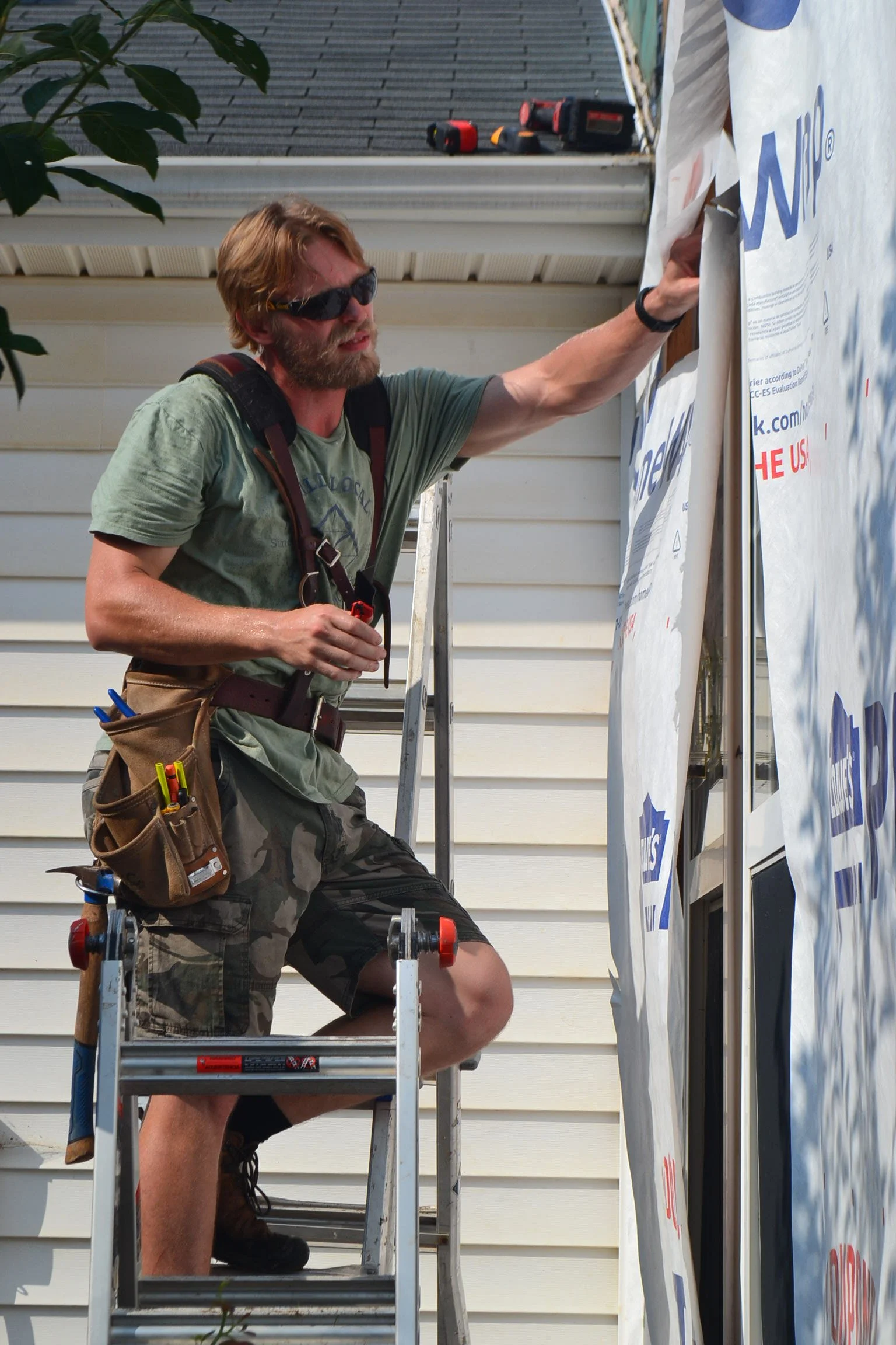 A man replacing the siding on a house for a repair company in Greenville, South Carolina. He is wearing a tool belt and sunglasses.