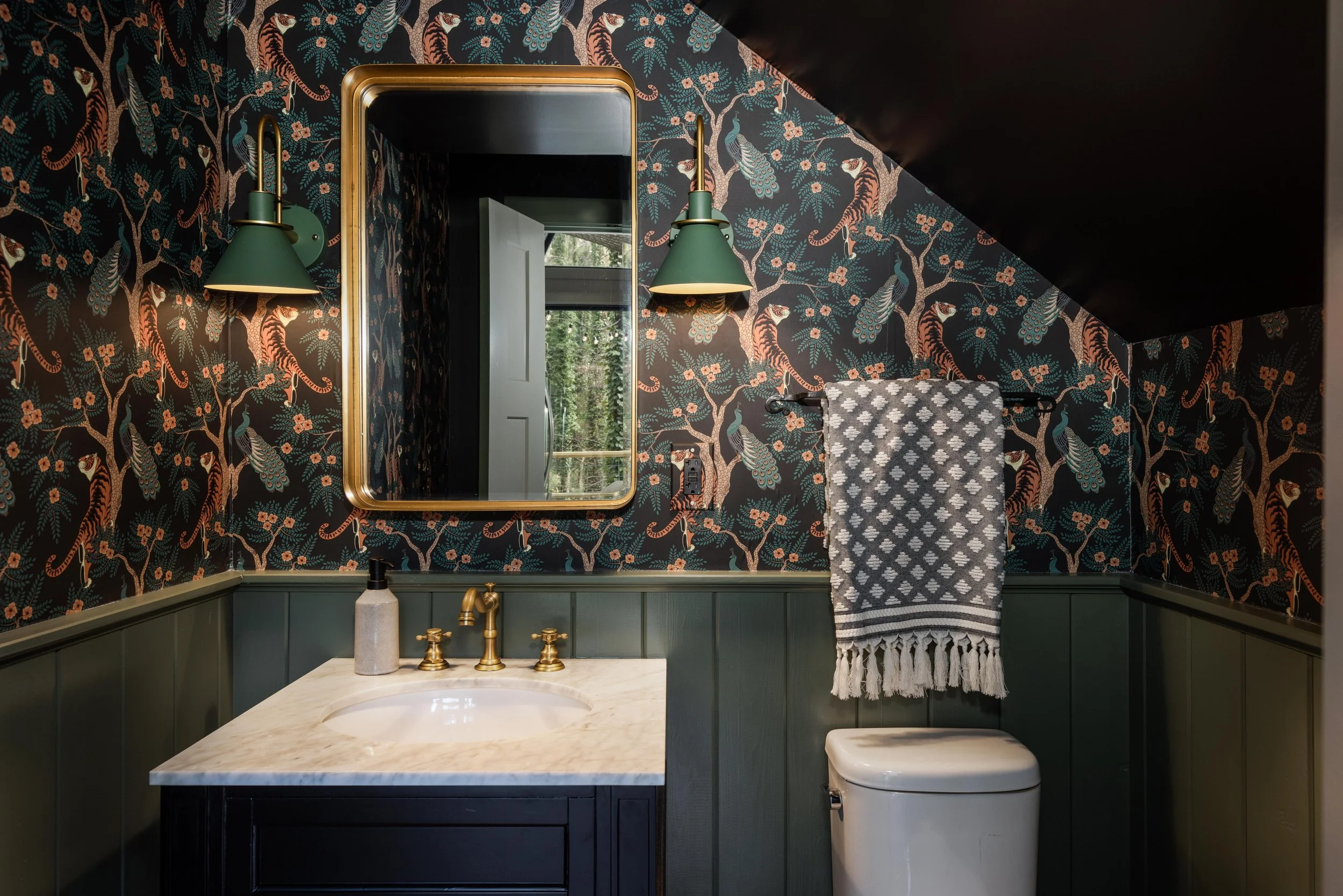 A bathroom with dark tiger print wallpaper, a mirror, green wall-mounted lights, a marble countertop with a sink, and green cabinetry. There is a window reflecting trees outside and a towel hanging on a rack.