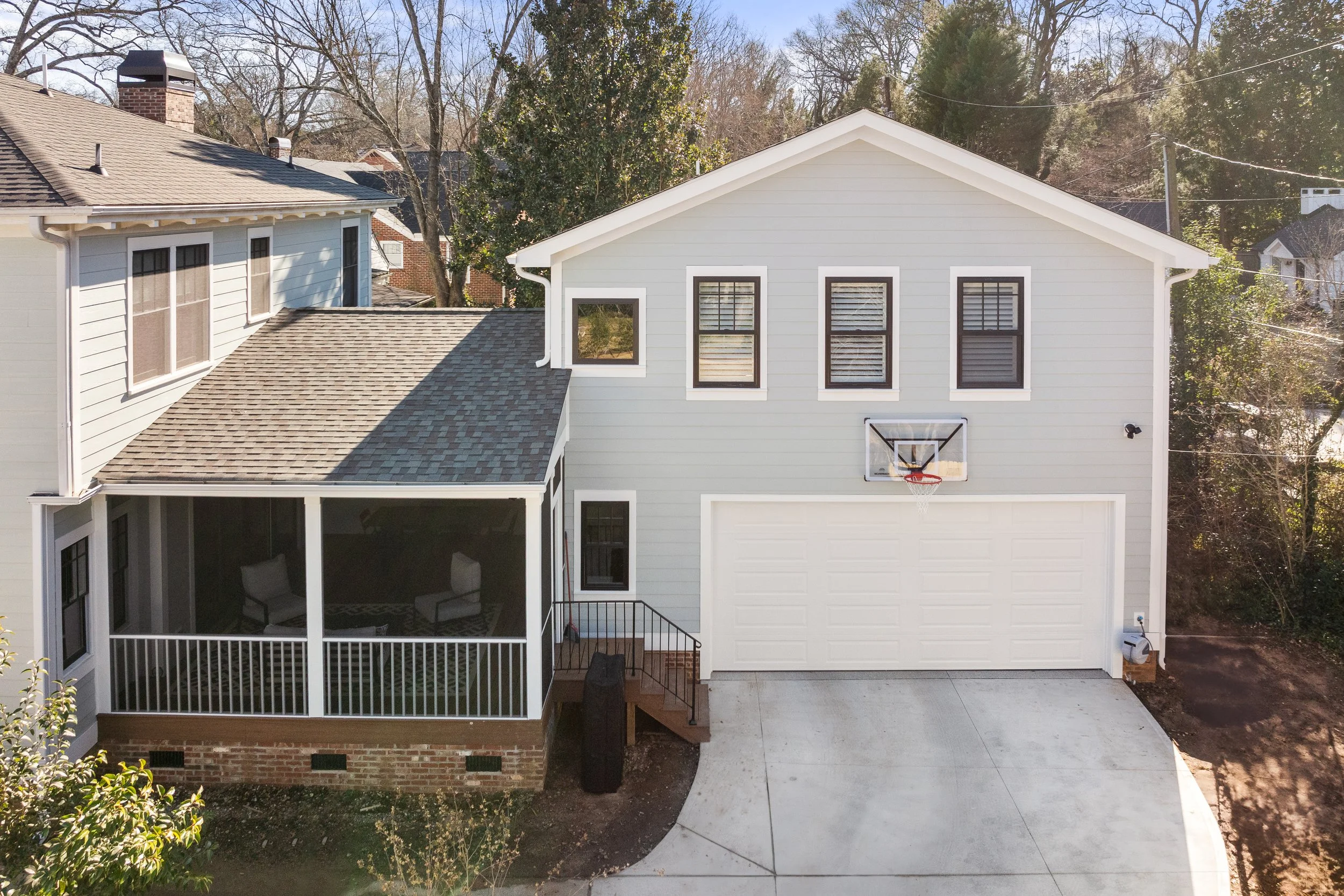 A modern two-story house with a white exterior, multiple windows with black frames, a garage, and a front porch. There is a basketball hoop above the driveway, and trees surrounding the house.