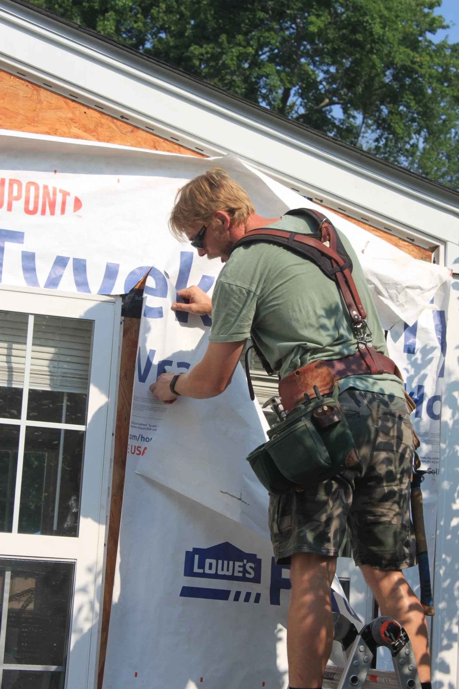 A man working on a home exterior near a Lowes banner, wearing camouflage shorts and a green t-shirt, on a ladder during daytime.