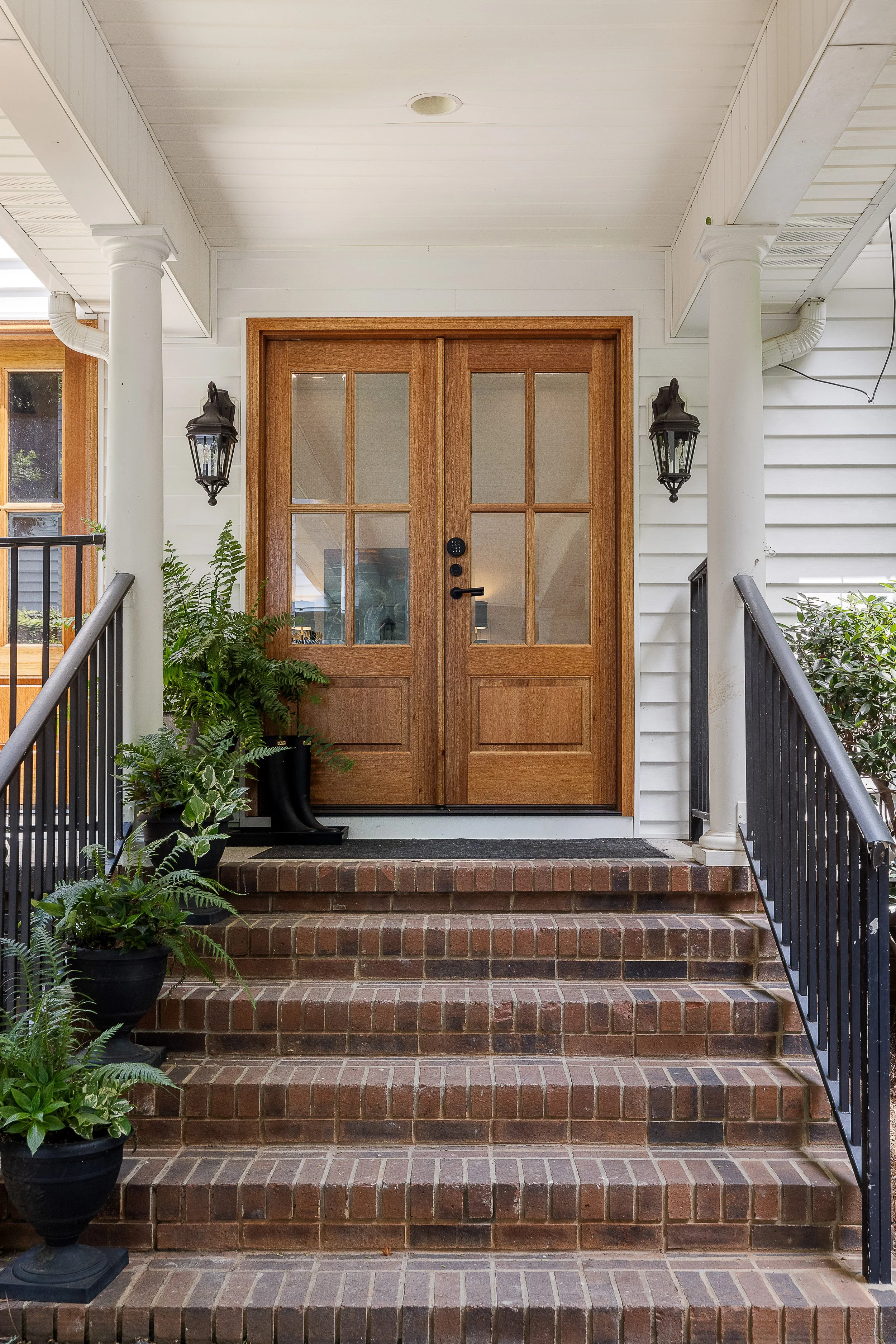 Front porch of a house with brick steps, white siding, and a wooden door with glass panes. Black metal railings on each side of the steps, potted plants on the left, and black outdoor wall lanterns on each side of the door.