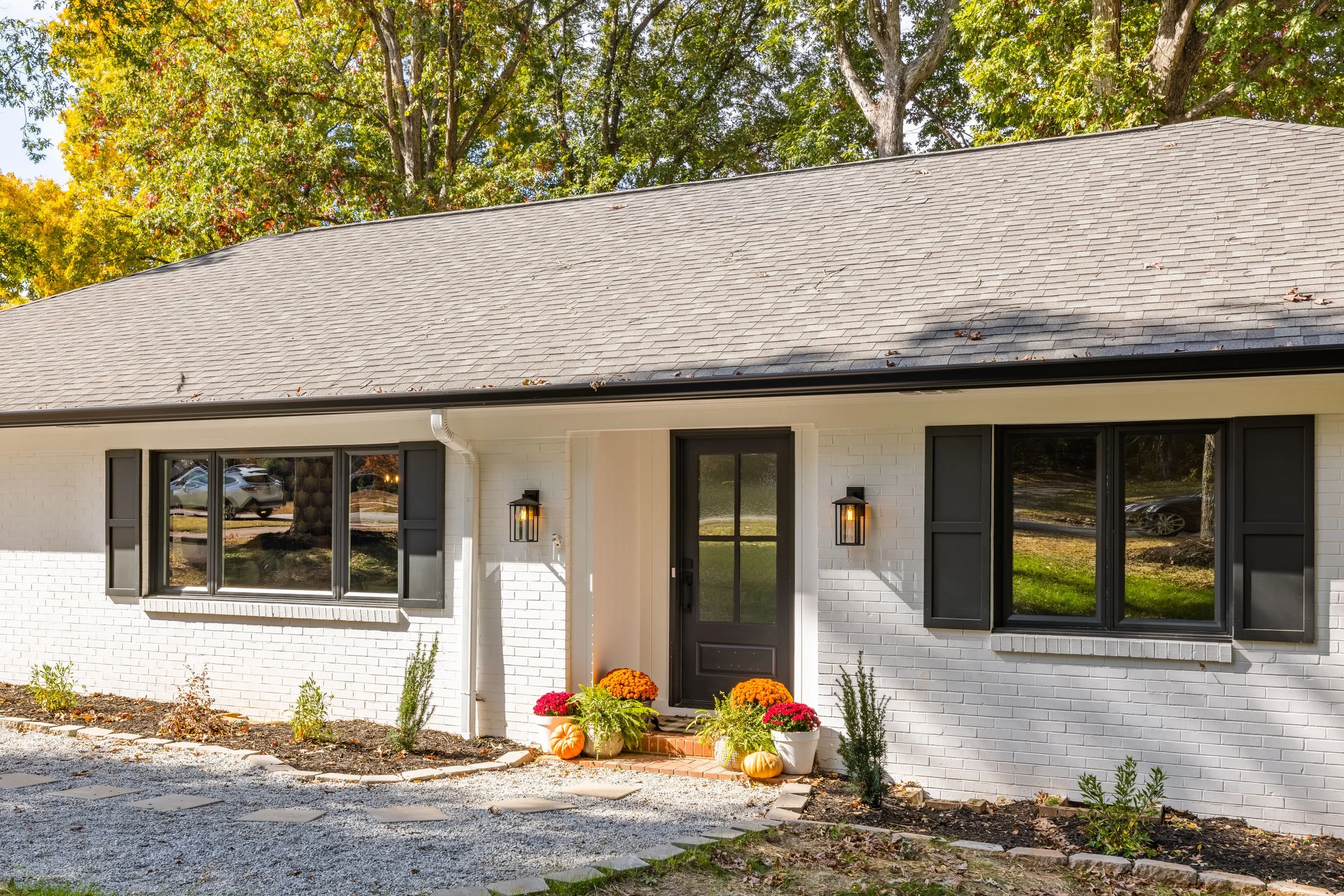 Front view of a white brick house with black window shutters and a black door, decorated with pumpkins and potted chrysanthemums, with large trees in the background.