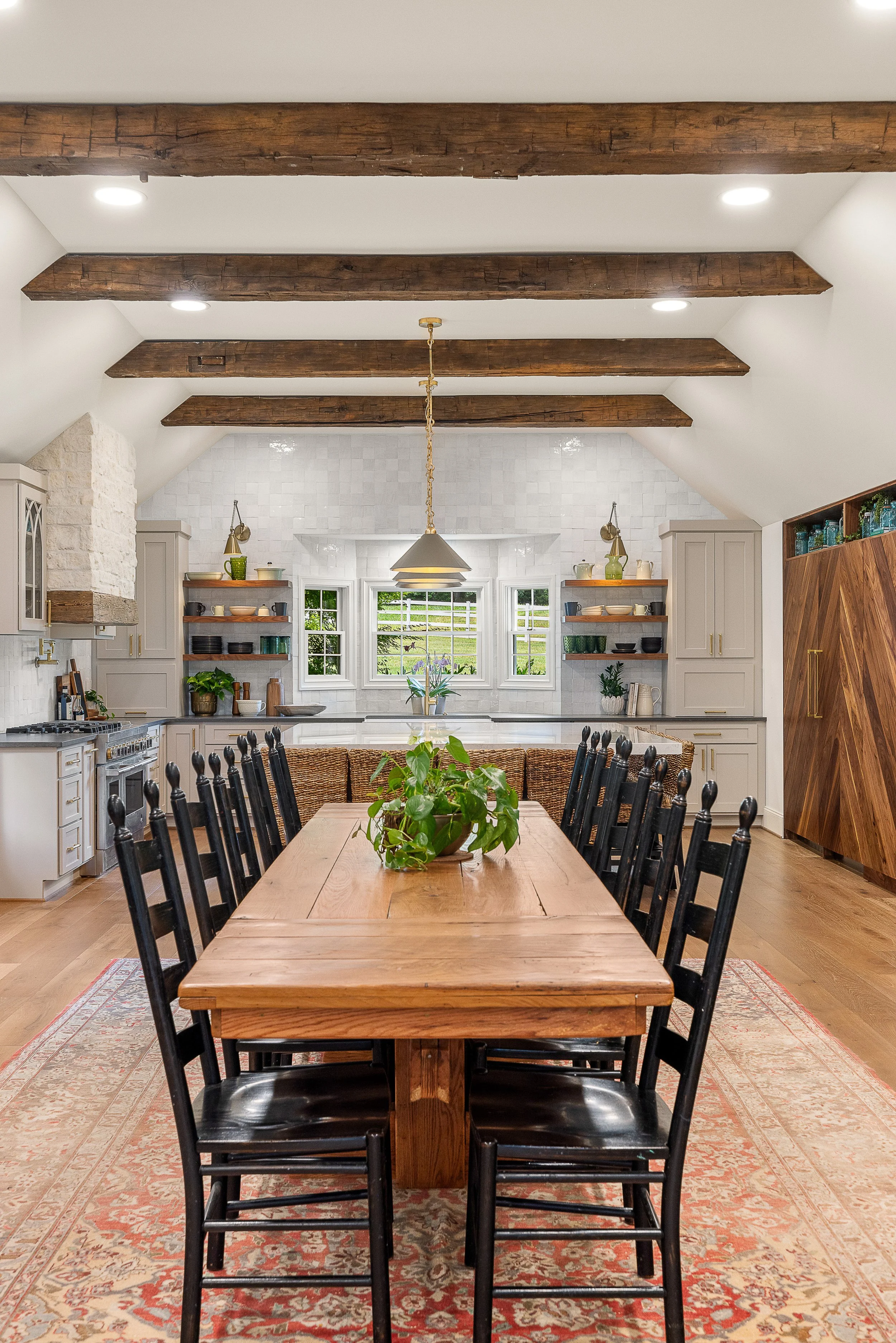 A spacious dining room with a long wooden table, black chairs, a decorative plant centerpiece, and a patterned rug. In the background are white kitchen cabinets, open shelves with dishes, and three windows showing a green outdoor landscape. The ceiling has wooden beams and recessed lighting.