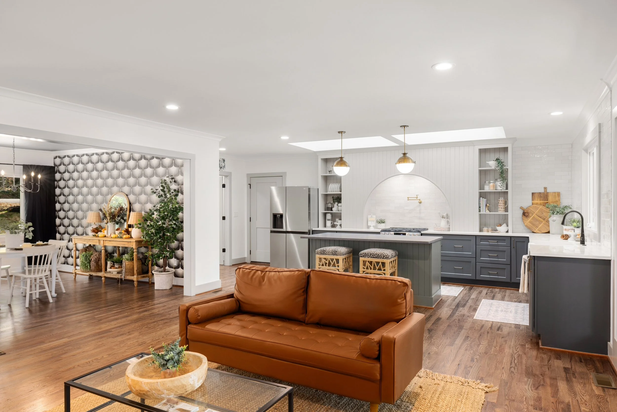 Open concept living and kitchen area with white walls, gray and white kitchen cabinets, a kitchen island, and dining area. Decor includes plants and a black and white wallpaper accent wall.