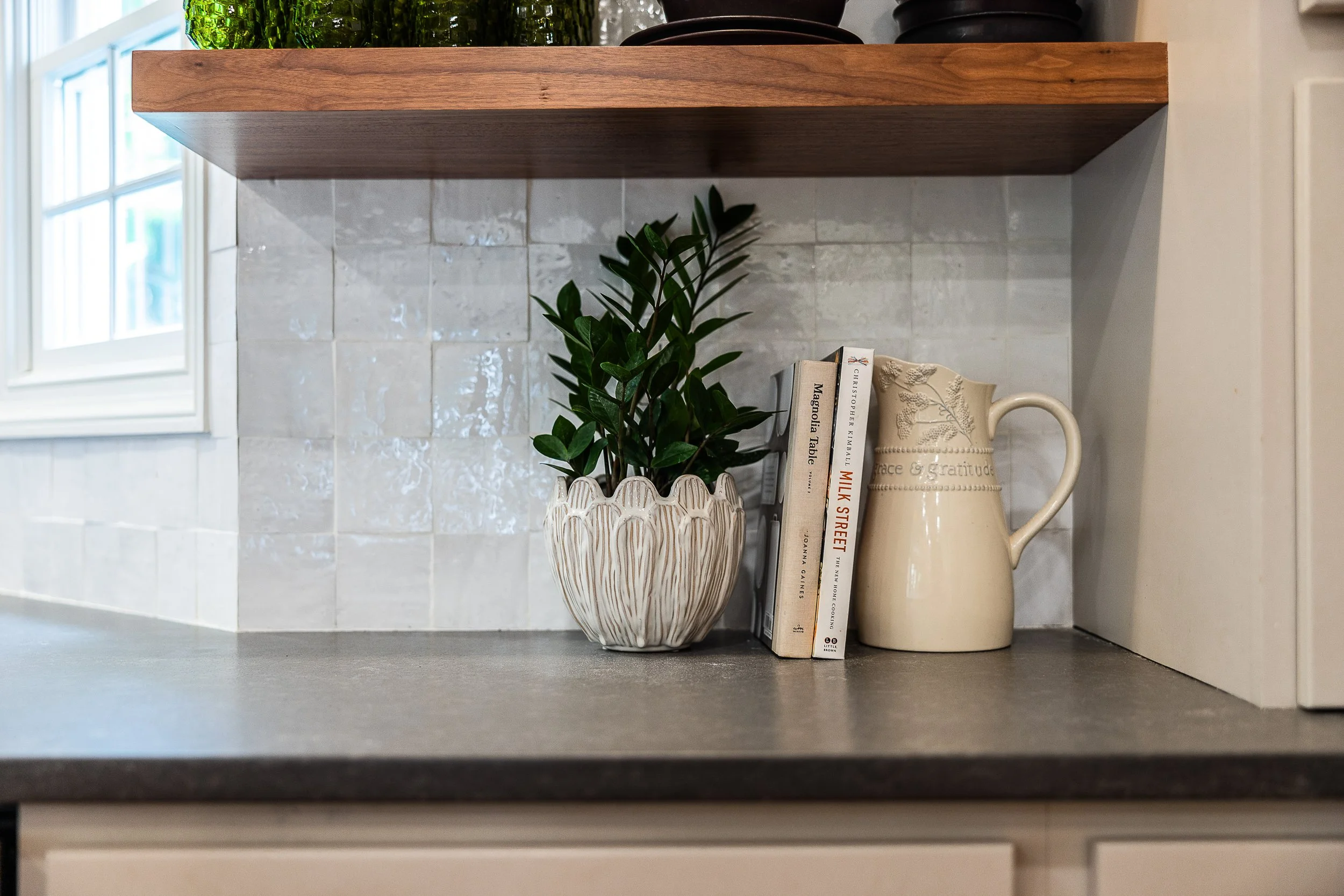 Decorative arrangement on a kitchen countertop including a white ceramic pot with leafy green plant, a small stack of cookbooks, and a cream-colored jug with embossed floral design, set against a tiled backsplash and partially visible window.