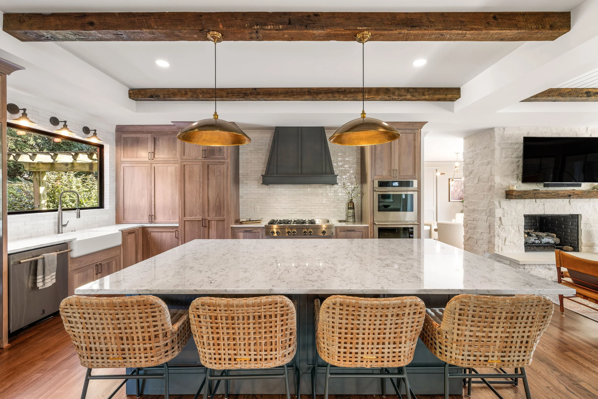 Modern kitchen with a large white marble island, woven barstools, wooden cabinetry, black range hood, brick backsplash, large window, and a stone fireplace with a mounted TV.