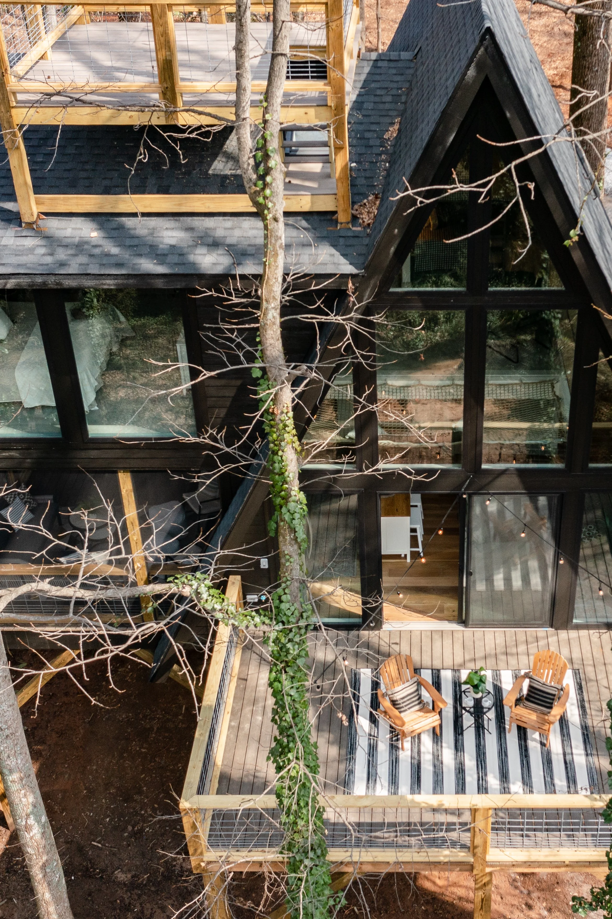 Bird's eye view of a modern house with black metal framing and large glass windows. The house has a deck with two wooden chairs and a small table. A new wooden extension is under construction with framing and railing, and sparse trees or branches are visible in the foreground.
