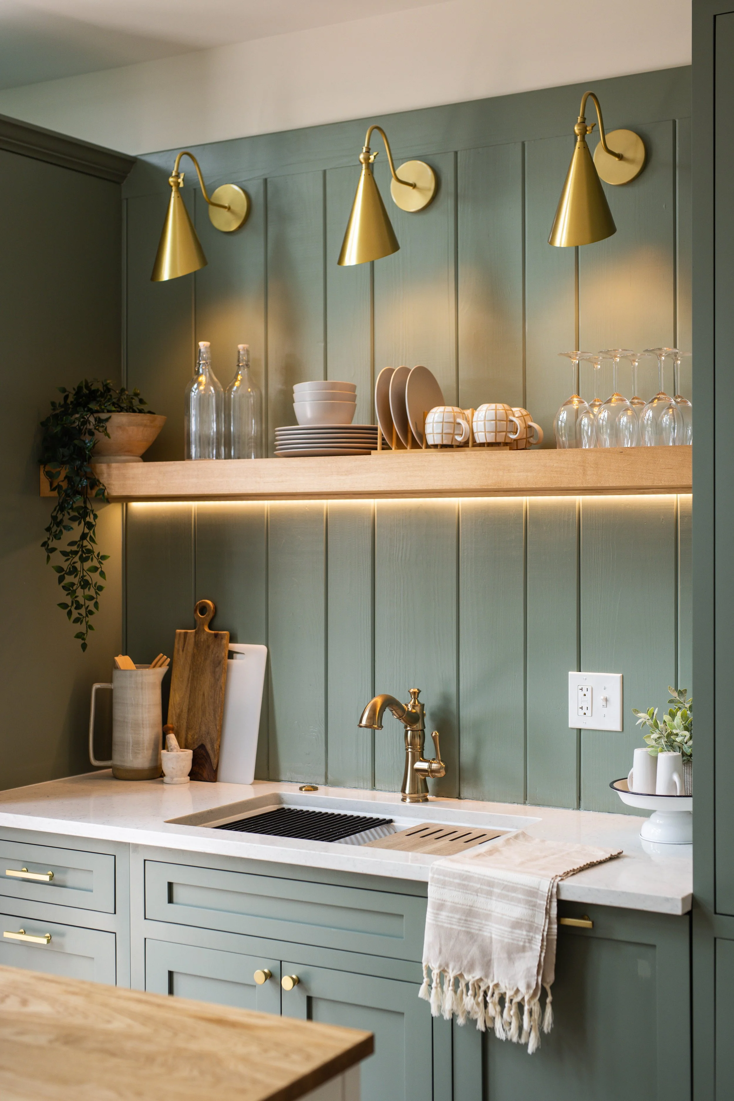 A kitchen with a white countertop, green cabinets, and a white backsplash. There are three gold wall-mounted lamps above the counter. On the counter, there are glass bottles, white bowls, beige plates, and mugs. A plant is on a shelf, and a private faucet is visible with a beige towel hanging on the cabinet handle.