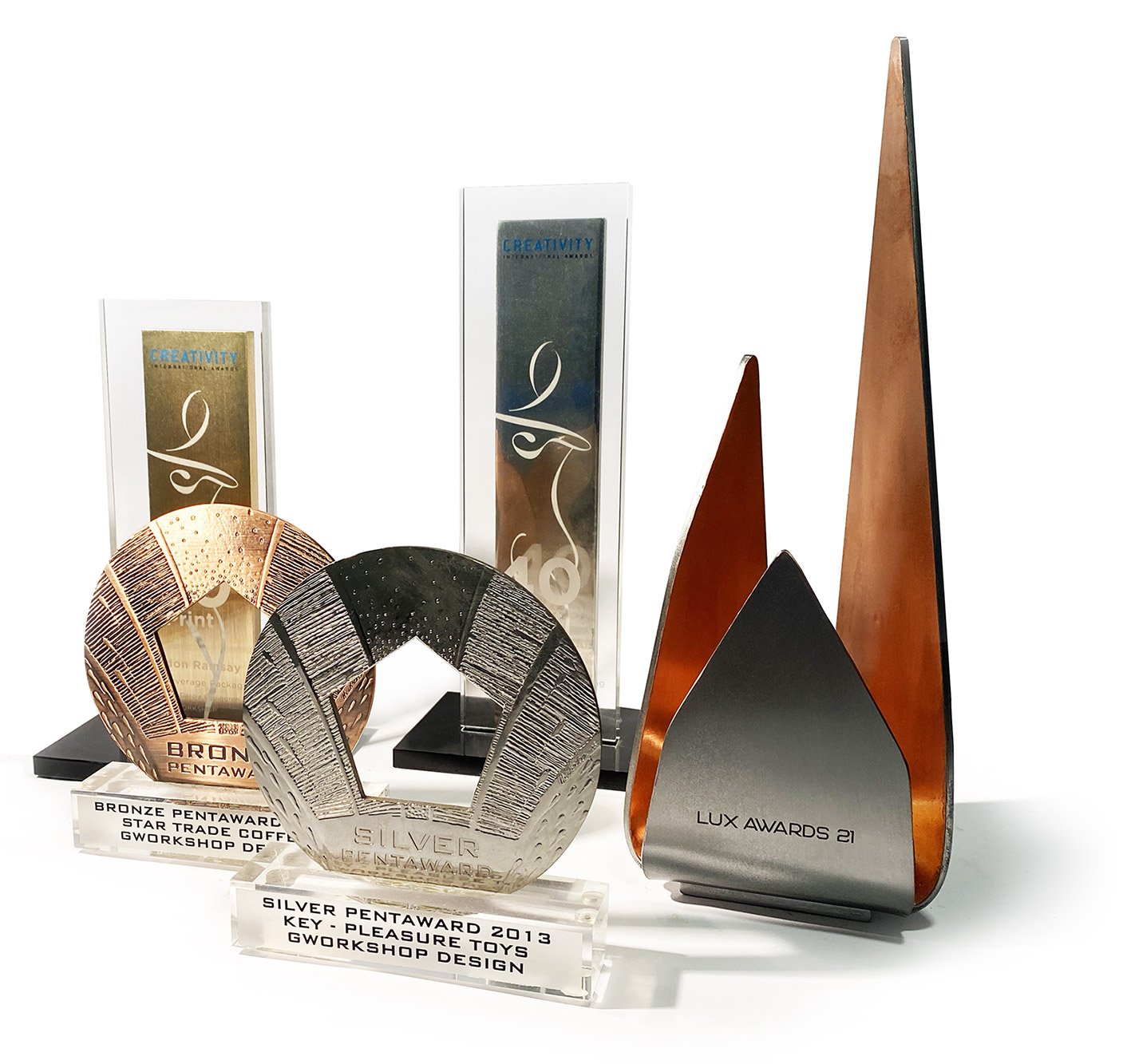 Premios y trofeos de diseño en varias formas y colores, incluyendo trofeos de metal y acrílicos, con inscripciones como 'Silver Pentaward 2013' y 'Lux Awards 21.'