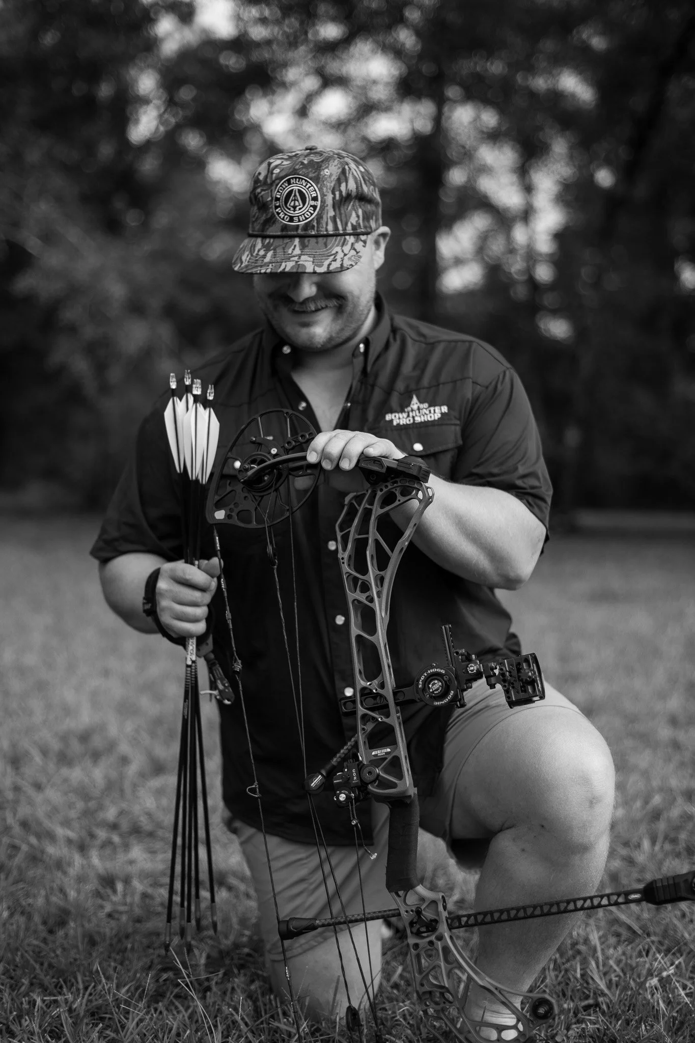 A man kneeling on the grass outdoors, holding a modern compound bow with a sight and stabilizer, wearing a camouflage cap and a button-up shirt with a logo, smiling while looking down at the bow.