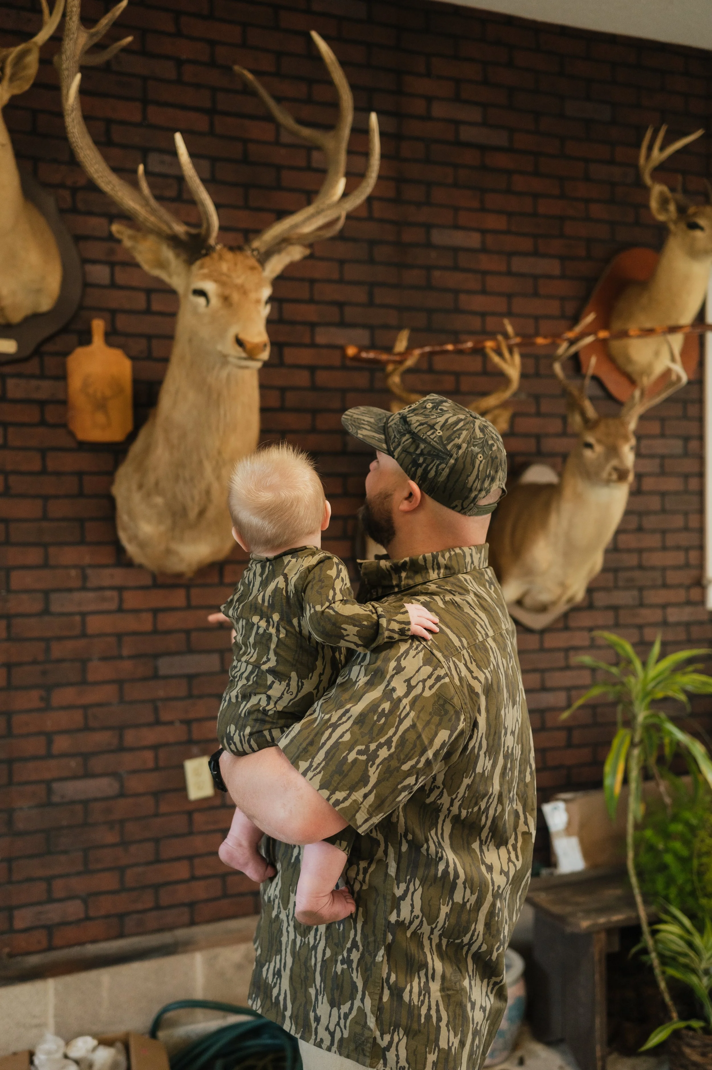 A man in camouflage clothing is holding a young child who is also dressed in camouflage clothing, standing in front of a wall with mounted deer heads and antlers in an indoor setting.