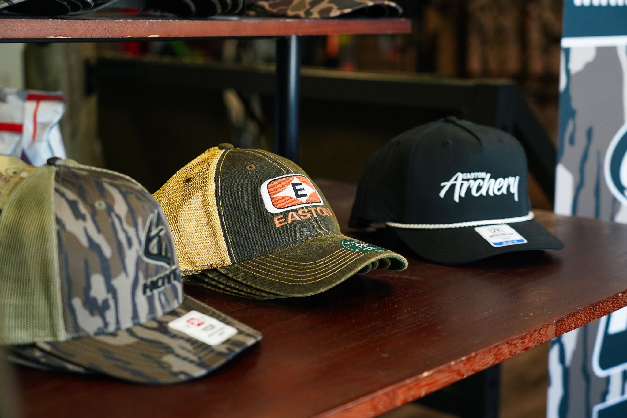 Several baseball caps displayed on a wooden table, with a brown cap featuring the Easton logo, a black cap with 'Easton Archery' written on it, and other camouflage and patterned caps.