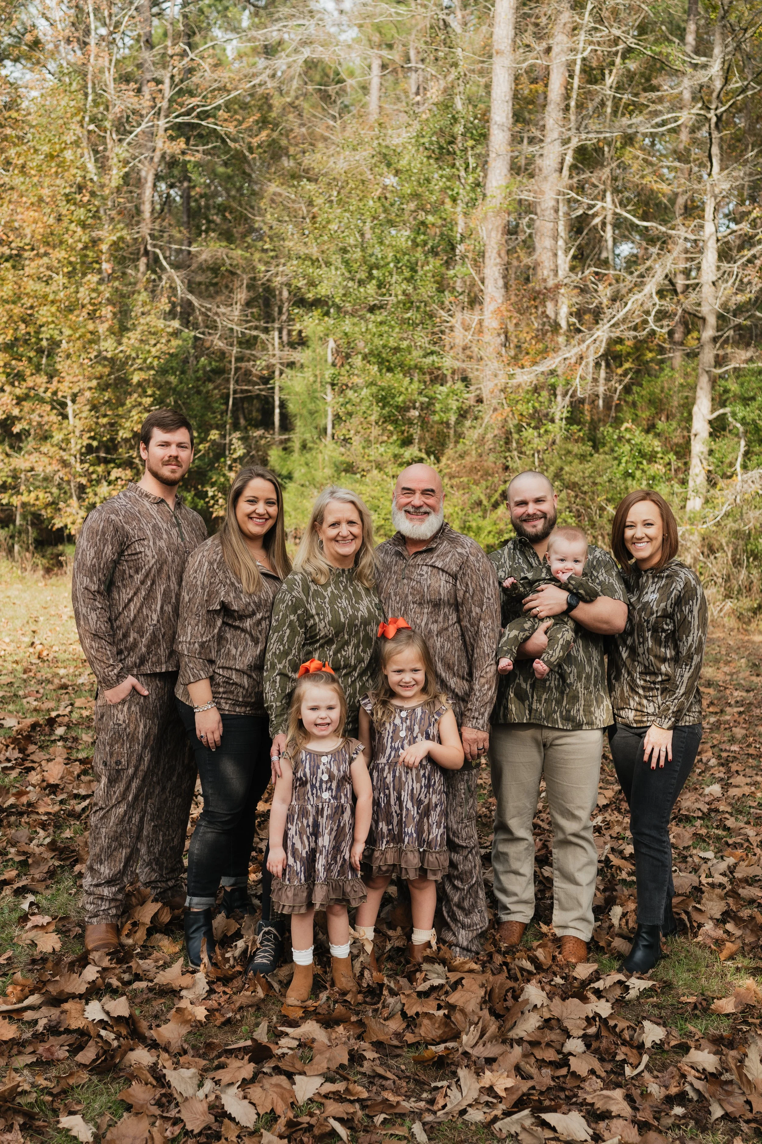 Group of six adults and four children standing outdoors among fallen autumn leaves with a forest in the background, wearing matching camouflage-patterned clothing.
