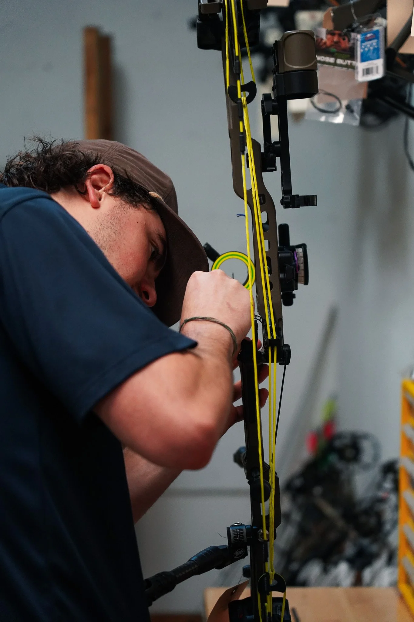 A man wearing a brown cap and glasses is aiming a compound bow with yellow strings, focusing on the target.
