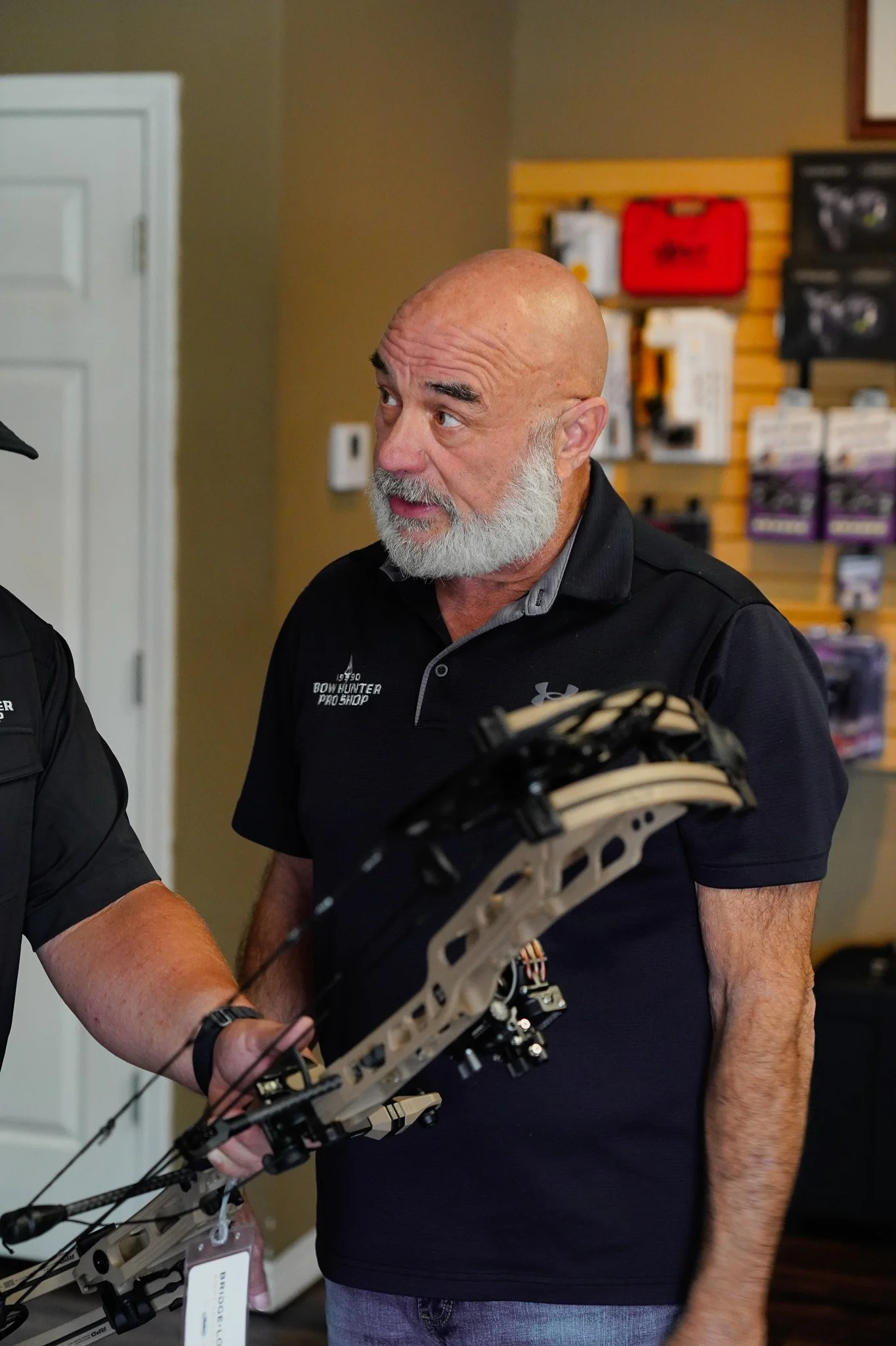 A man with a gray beard wearing a black polo shirt holding a compound bow with a scope and bow sight in an indoor store.