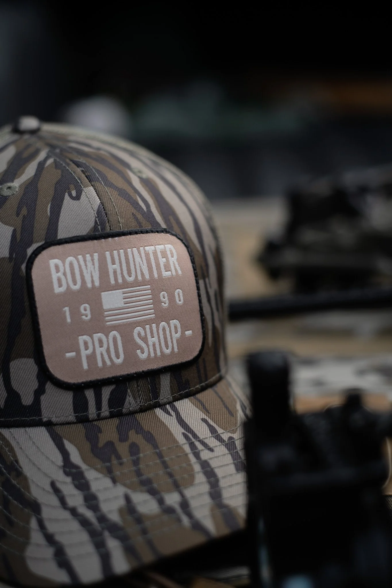 Camouflage baseball cap with a patch reading "Bow Hunter" and an American flag, placed on a surface with blurred objects in the background.