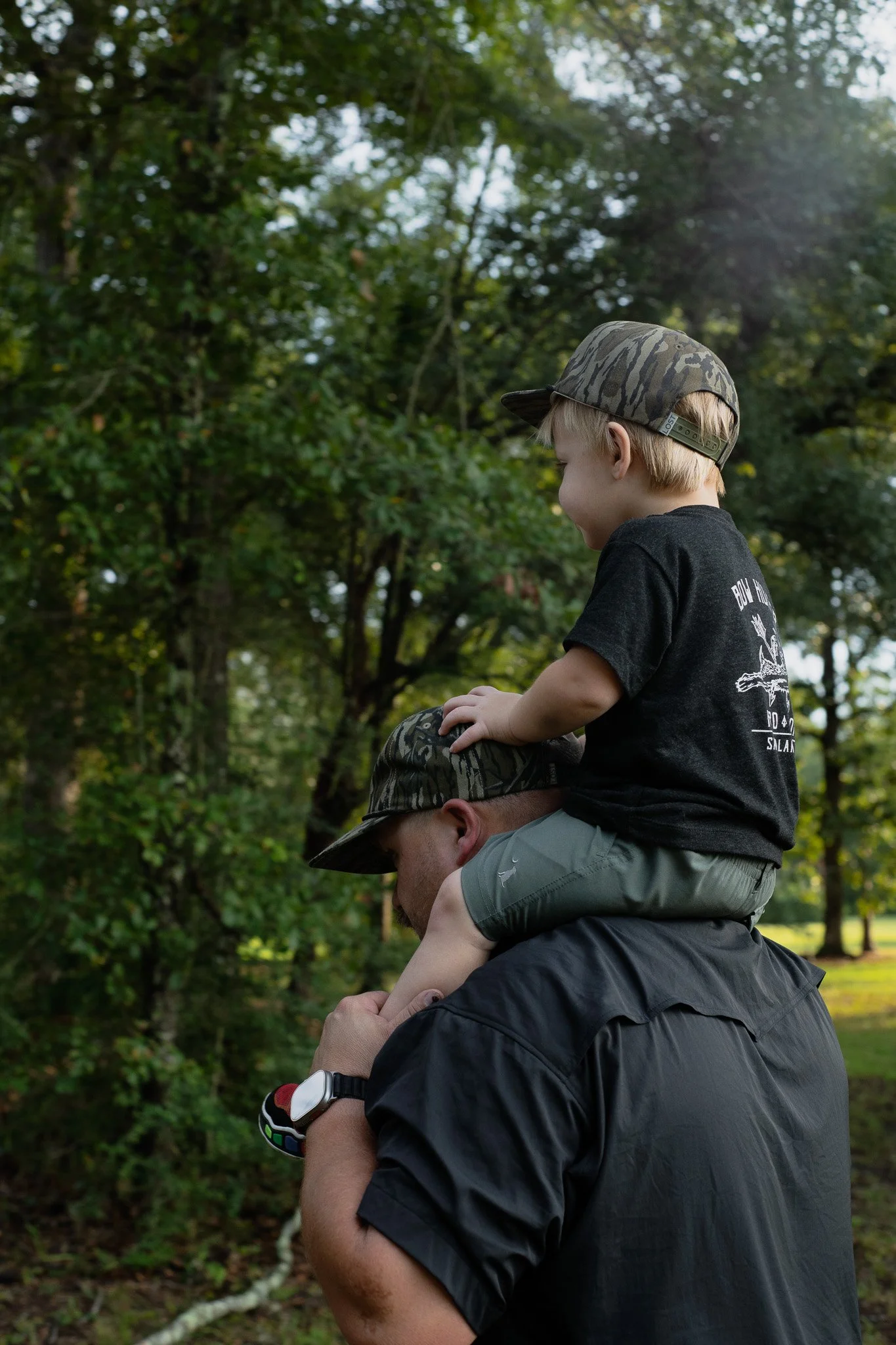 A man is carrying a young boy on his shoulders outdoors in a park with trees in the background. The boy is wearing a camouflage cap and black T-shirt, and the man is wearing a dark shirt and a watch.