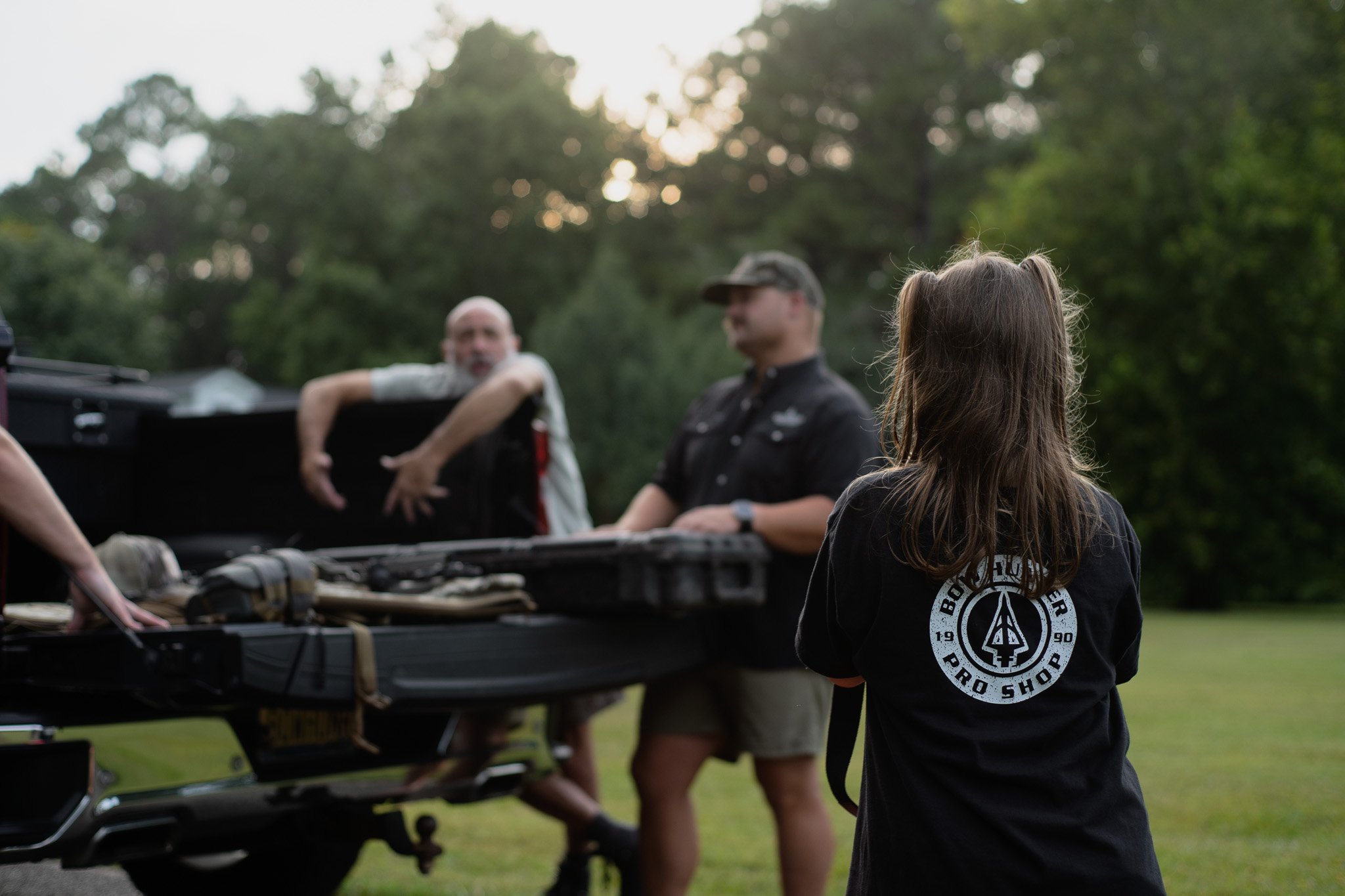 Young girl with long hair and a logo on her black shirt watching men working on a car engine outdoors during daytime.