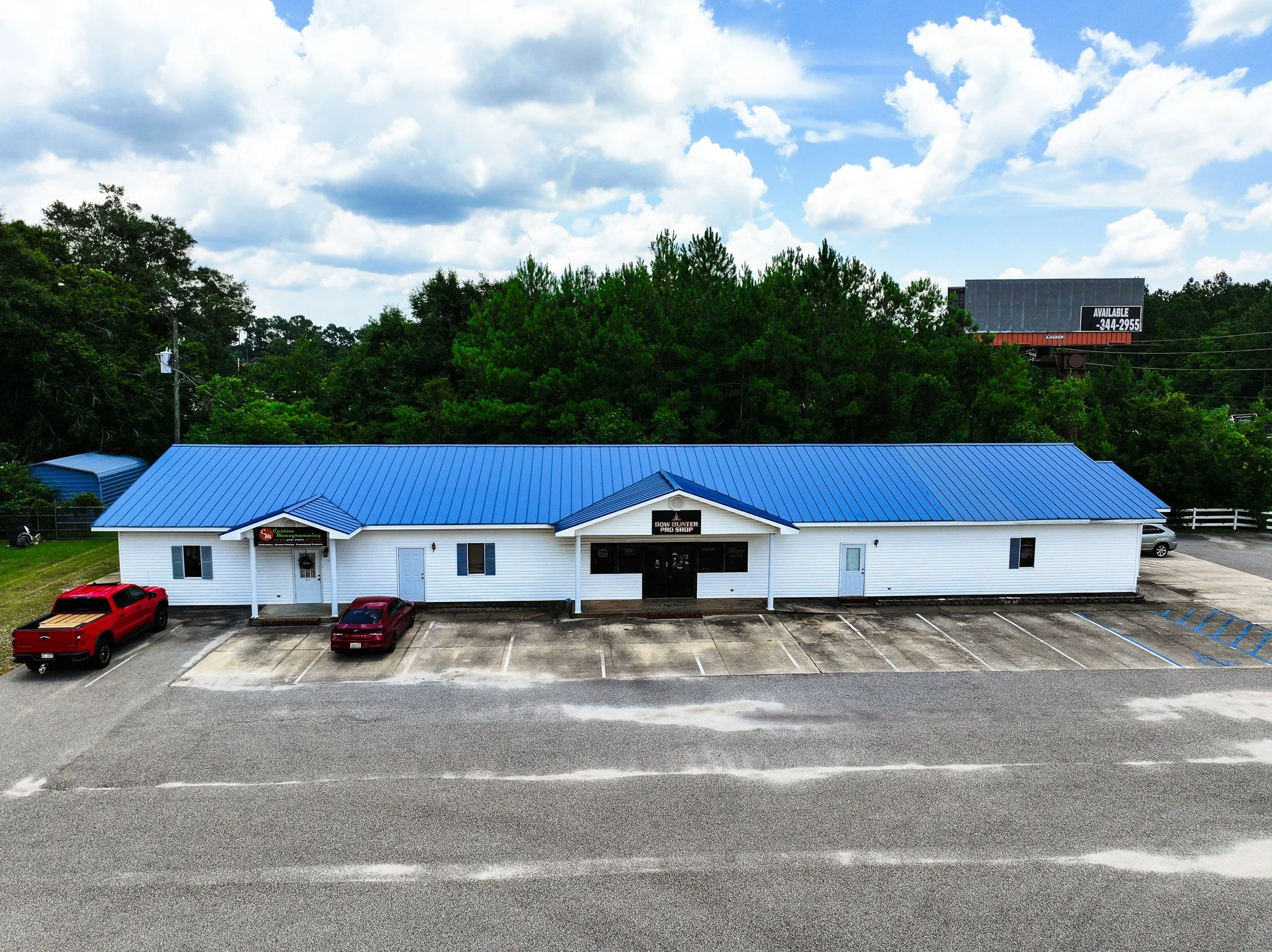 Empty parking lot in front of a white building with a blue metal roof, surrounded by trees under a partly cloudy sky.
