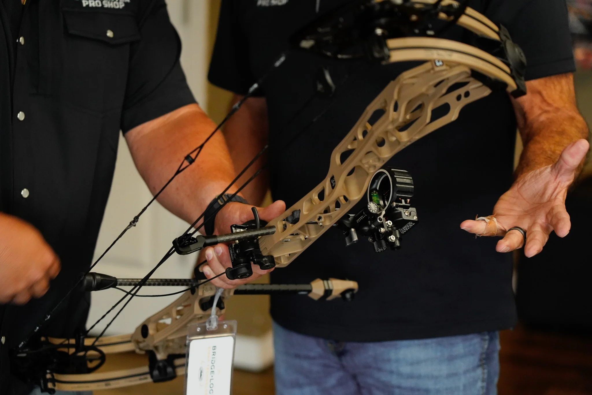 Two people are holding and examining a tan-colored compound bow with black components at an indoor event or store.