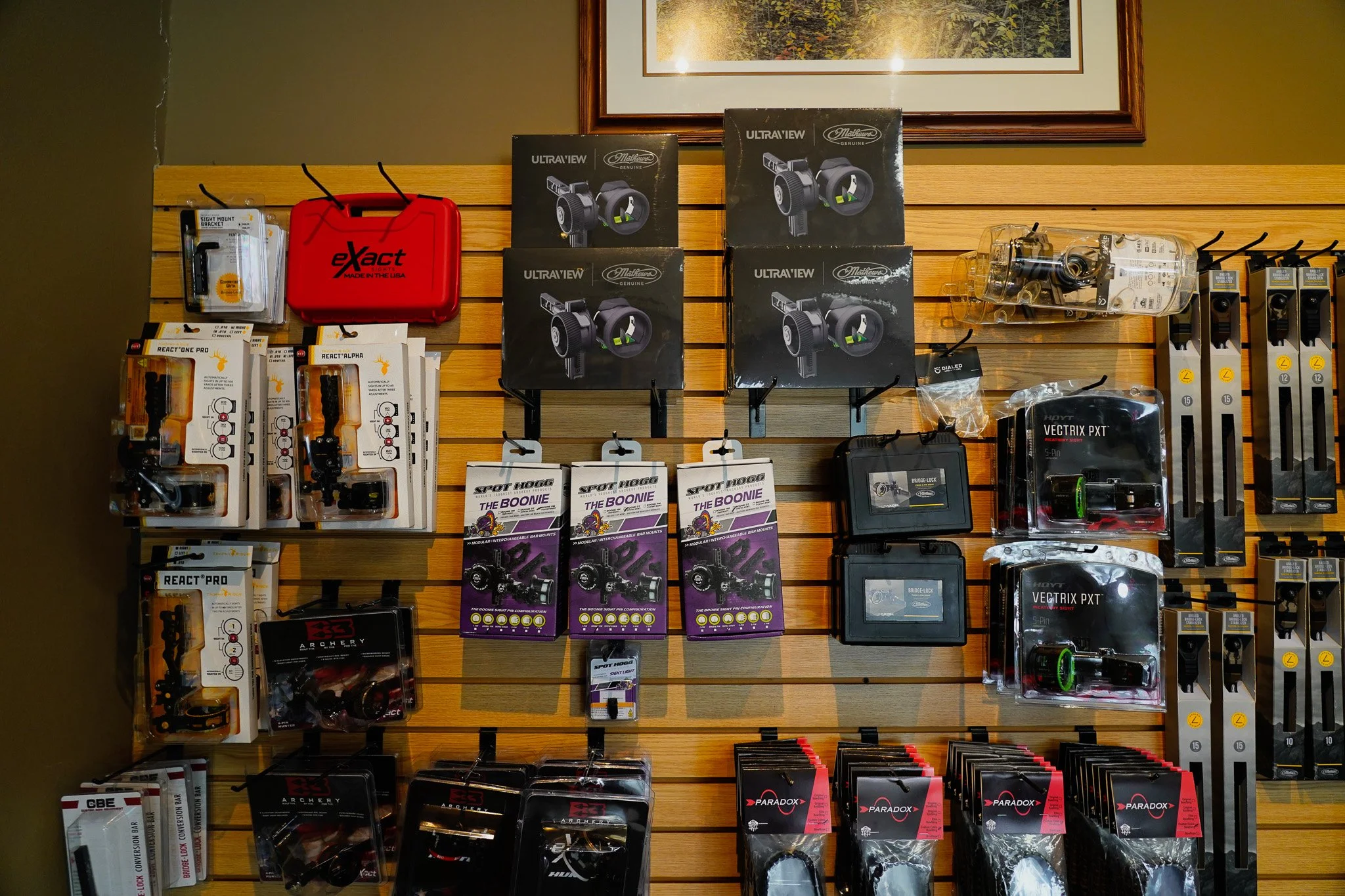 A wooden display wall with various archery equipment and accessories, including sight mount brackets, boxes of sight gear, arrow rest systems, and other archery tools and accessories, arranged in an organized manner.