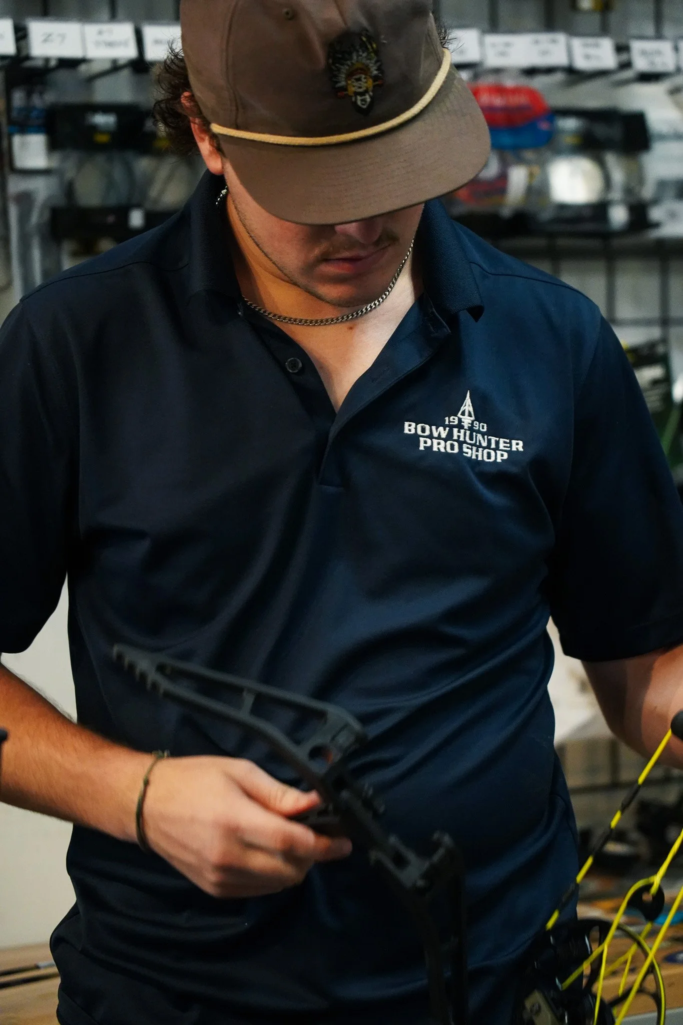 A person wearing a navy blue polo shirt with 'Bow Hunter Pro Shop' embroidered on it, a brown cap, and a silver chain necklace, holding a bow and working with yellow and black strings or wires in a workshop setting.