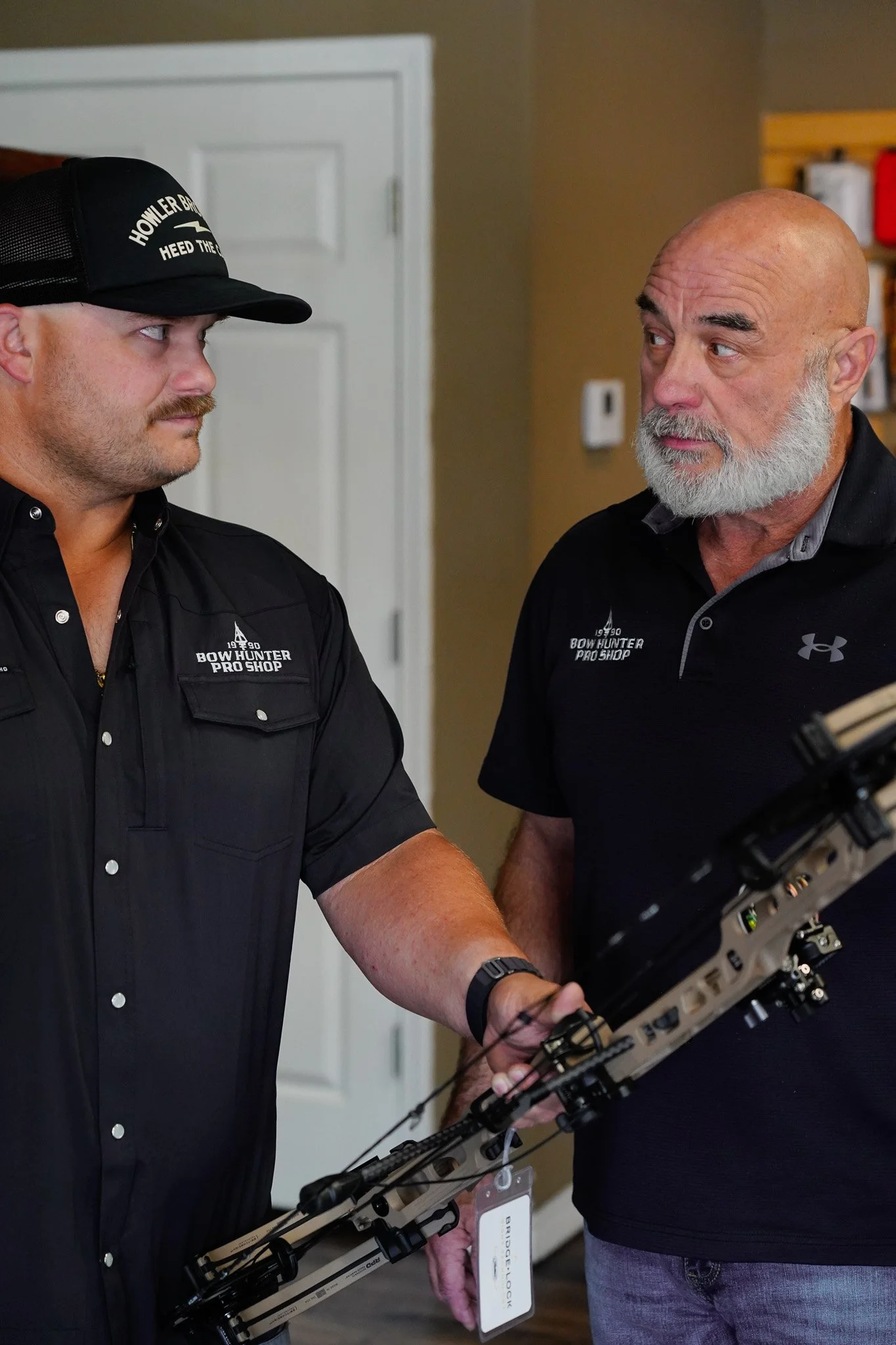 Two men are inspecting a crossbow. The man on the left is wearing a black shirt and cap, with a badge that reads 'Bow Hunter Pro Shop.' The man on the right has a grey beard, is bald, and is wearing a black Under Armour shirt. They are standing indoors, with a white door and a wall-mounted thermostat visible in the background.