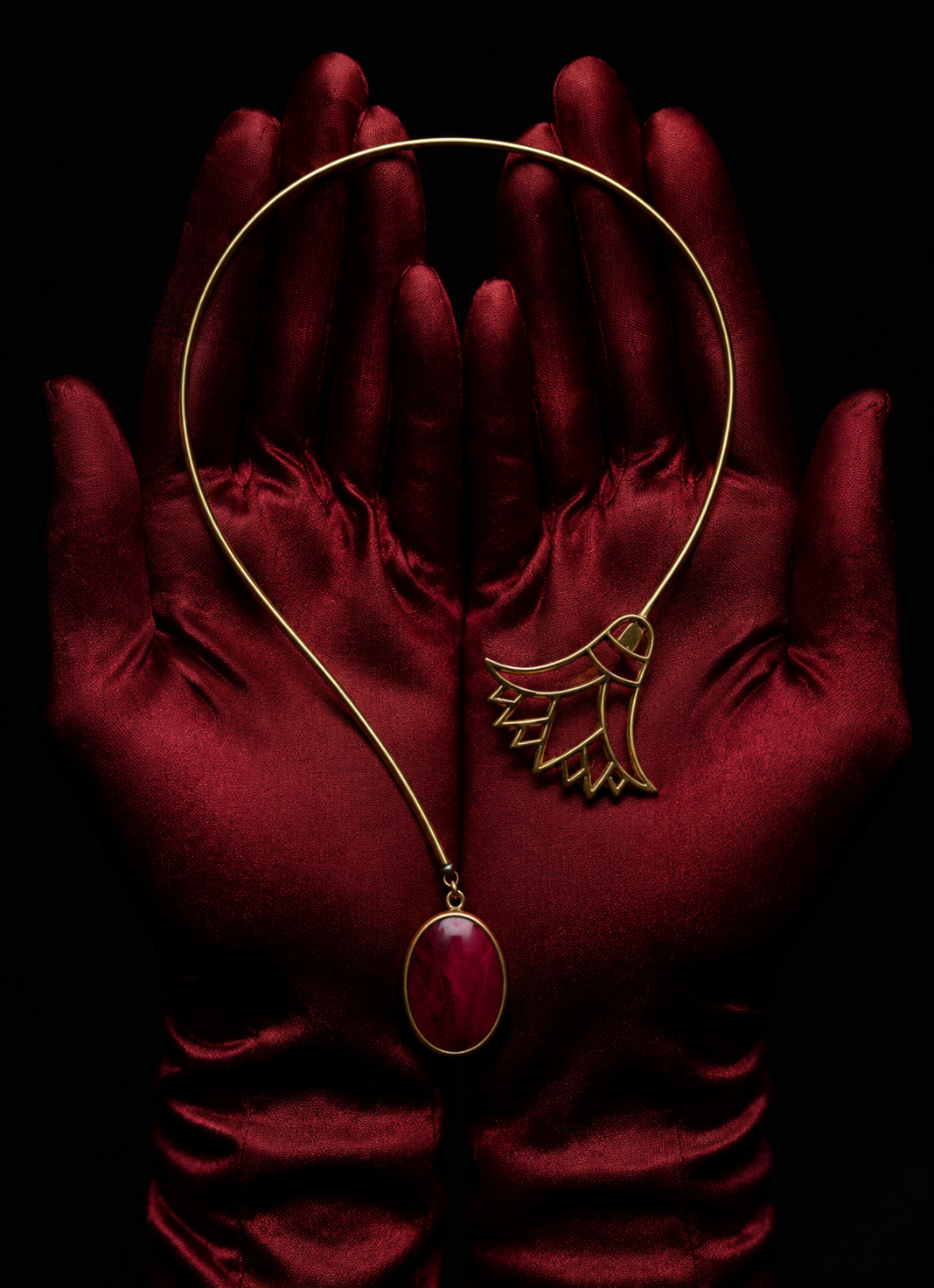 A pair of red satin gloves holding a gold necklace with a red oval pendant and a gold leaf-shaped design on a black background.